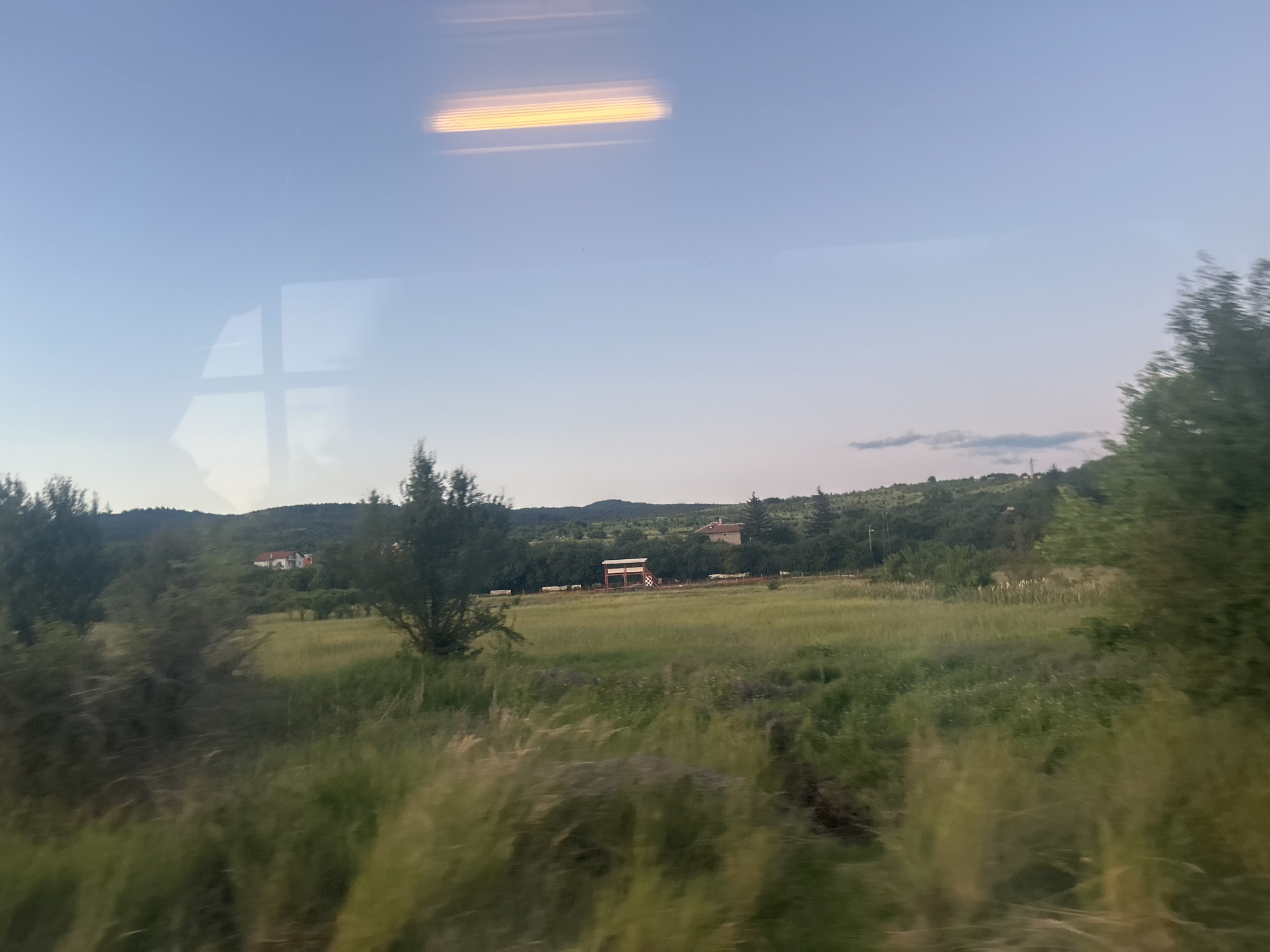 The image depicts a scenic countryside view, likely captured from a moving vehicle due to the motion blur in the foreground and reflections on the surface. The sky is a pale blue with a few faint clouds, and a bright, horizontal reflection of what appears to be lights is visible in the upper portion of the sky. Below the sky, rolling green hills covered in trees form the background. In the middle ground, a large, grassy field is dotted with trees and a few buildings. One prominent building is a red-roofed structure, possibly a house or a barn, situated on the hillside. There is also a small, elevated observation-like structure with a red roof in the middle of the field. The foreground is dominated by tall, blurry grass and vegetation, further emphasizing the sense of motion. The overall impression is of a tranquil, rural landscape.