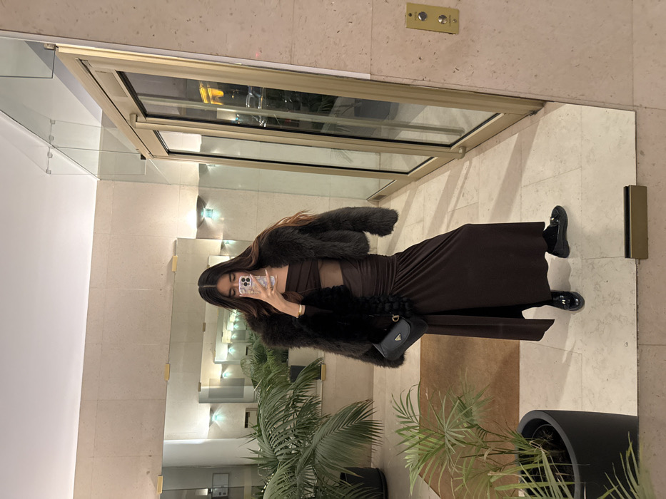 A woman is seen in a mirror selfie, wearing a brown dress with a midriff cutout, a fuzzy jacket, and black shoes. She is holding a phone to take the picture. The reflection shows a well-lit interior with architectural details and greenery, including potted plants near the base of the mirror. A switch panel is visible on the wall.