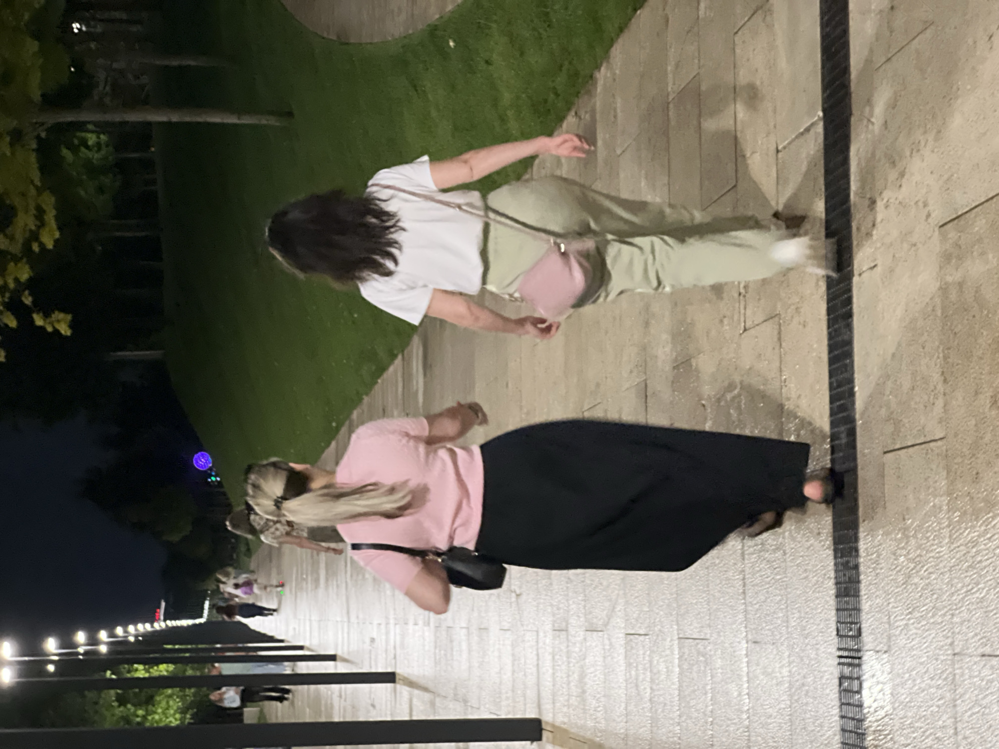Two women are walking away from the camera on a paved pathway in a park at night. The woman on the left has blonde hair in a ponytail and is wearing a pink t-shirt and a long black skirt. She is carrying a black handbag on her left shoulder. The woman on the right has dark hair and is wearing a white t-shirt and light green pants. She is carrying a light pink crossbody bag. The pathway is illuminated by streetlights. In the background, there is a grassy slope with trees and more streetlights. There are other people walking in the distance, some of whom appear to be children. The image was taken in Krasnodar, Russia.