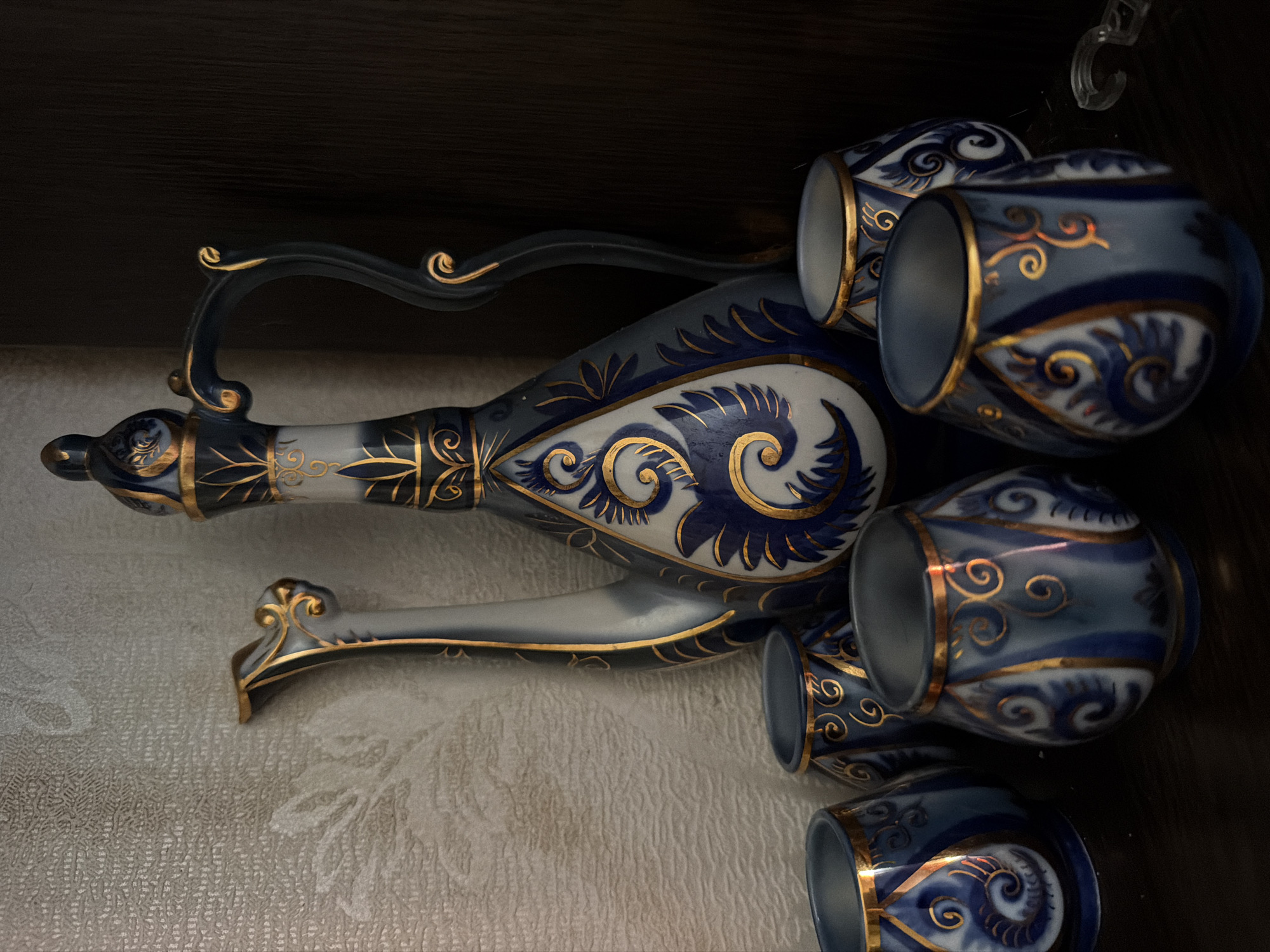 A decorative ceramic set, consisting of a tall pitcher with a curved handle and five smaller cups, is displayed on a dark wooden shelf against a textured, light-colored wall. The pitcher and cups are adorned with intricate blue and gold patterns, featuring swirling designs and floral motifs. The pitcher has a slender neck and a spout, while the cups are bulbous with wide openings. The objects appear to be in good condition, with no visible damage. The lighting in the scene is dim, casting shadows and highlighting the decorative elements of the ceramics. The image is a still life composition. The location in the city of Shepsi, Russia is not ascertainable from the image.