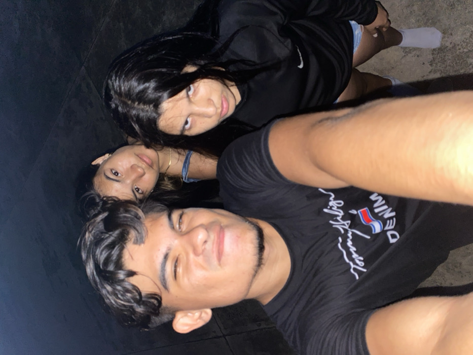 Three young people pose for a selfie at night. A young man wearing a black Tommy Hilfiger shirt is in the foreground. Behind him and slightly to the left is a woman with dark hair, wearing a light blue top. A third young woman, wearing a black Nike top and white socks, is positioned to the upper left of the frame. The background consists of dark, indistinct walls or ground.