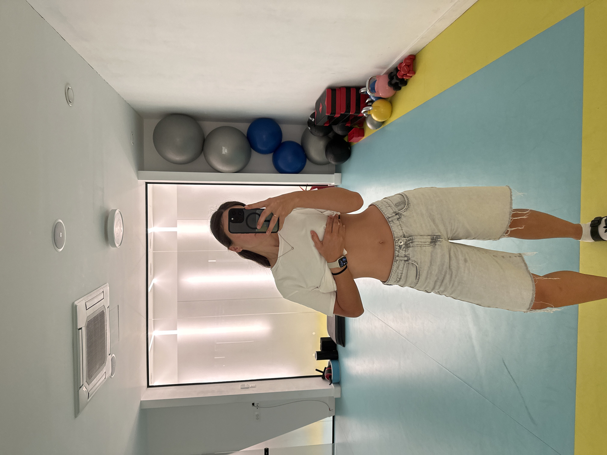 The image shows a woman taking a mirror selfie in what appears to be a gym or fitness studio. She is wearing a white t-shirt and light-colored denim shorts. The studio has light blue and yellow flooring. In the background, there are several exercise balls and some weightlifting equipment on a shelf. The walls are light blue and white. There is also a large air conditioning unit mounted on the wall. The lighting in the room is bright, suggesting it is daytime. The woman is holding her phone to take the picture, and her expression is neutral. There is no visible text in the image. The scene suggests a casual workout session. The location is Boreti, Montenegro.