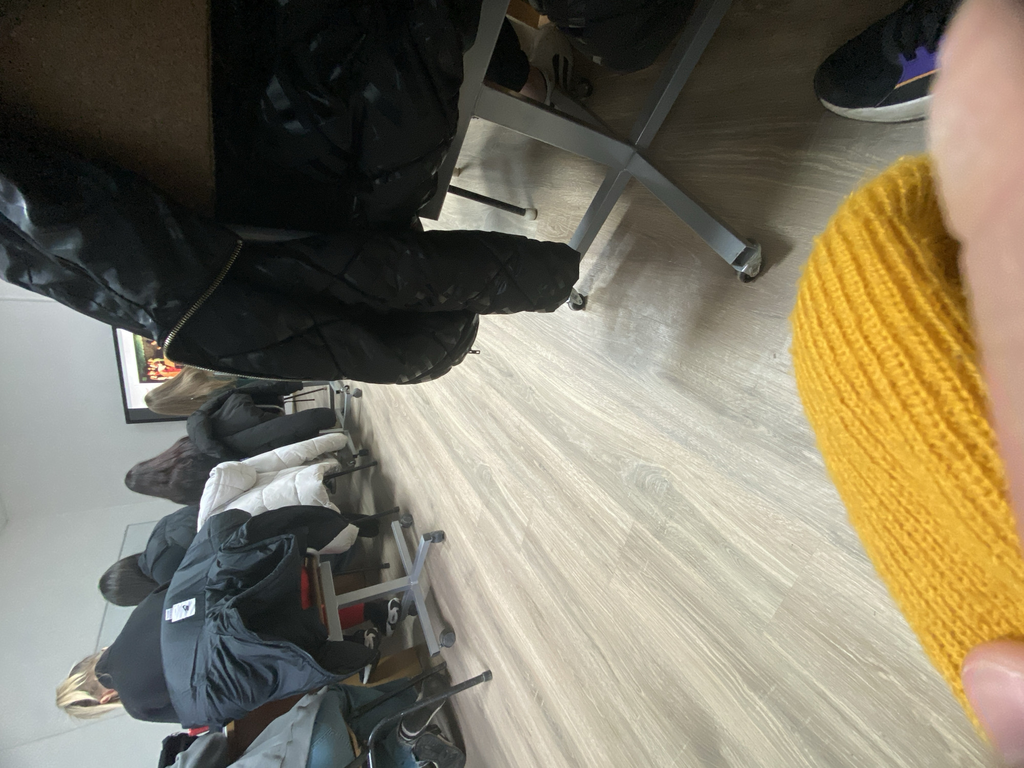 The image depicts a room with a light-colored, wood-grained floor. Several coats of varying colors (black, white, brown) are hanging on the backs of chairs that are lined up against the wall. Each chair is a standard office chair with a metal base and casters. In the upper right corner, a black sneaker with a purple accent and a yellow knitted object are visible. A partial view of a person is on the left side of the photo. The specific location in the city of Troyan, Bulgaria, is not discernible from the image. The photo contains chairs, jackets and people. The people are hanging jackets on the backs of the chairs.