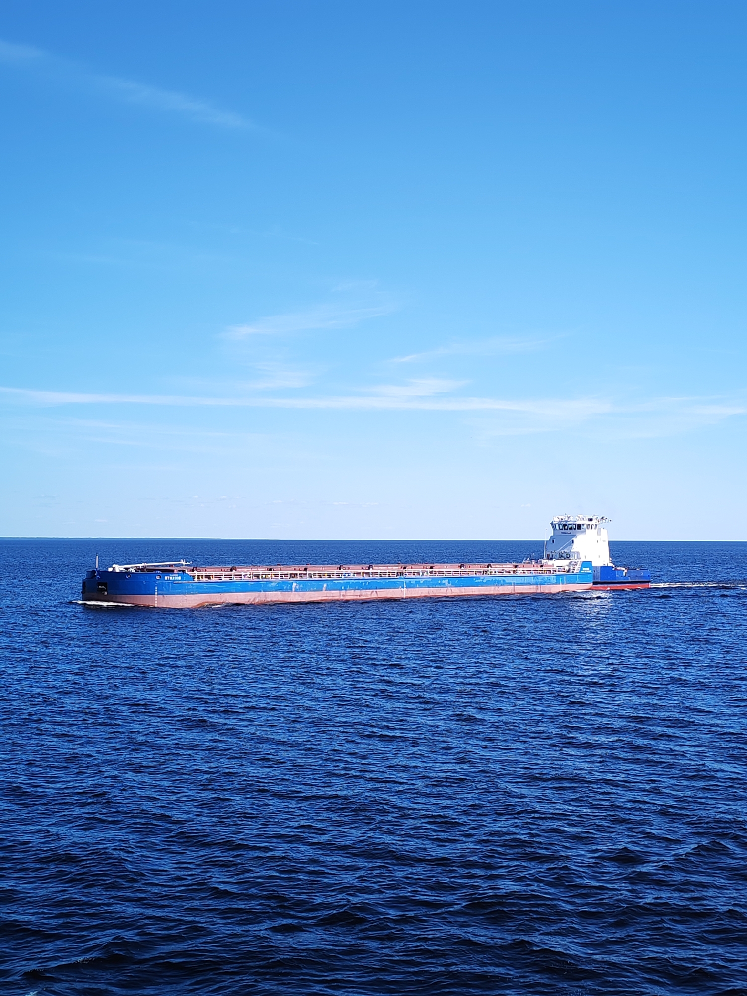 This image captures a cargo vessel, identified by the hull number "МУ 87368" and IMO number "7269046" (Rybinskaya-600), navigating a wide expanse of water under a clear blue sky, likely on the Rybinsk Reservoir near Мякса, Russia.
The vessel is a long, low-profile ship with a blue hull, a reddish-brown waterline, and a white superstructure located at its aft. It is positioned roughly in the center of the frame, moving from left to right, leaving a discernible white wake trailing behind its stern. The deck features white railings, and the cargo holds appear covered or empty.
The water is a deep cerulean blue, covered with numerous small, wind-induced ripples and waves, indicating light surface agitation. The horizon line is flat and clearly defined, separating the water from the sky. The sky above is a bright, uniform cerulean blue, with only faint, wispy white clouds scattered sparsely in the upper portion. The scene is devoid of other vessels, people, or visible landmasses in the immediate vicinity of the ship, portraying an open-water environment under sunny conditions.