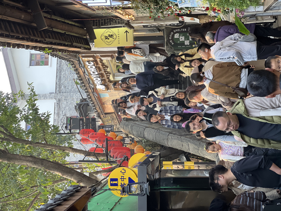This image depicts a crowded, multi-level pedestrian street and staircase in a tourist area within Yuzhong District, Chongqing, China. The scene is illuminated by natural daylight, likely late afternoon, casting warm tones.
The central feature is a wide, upward-sloping stone staircase, densely populated with approximately 50-60 individuals of varying ages, predominantly adults, engaged in pedestrian movement, ascending and descending. Most individuals are dressed in casual, layered attire.
On the left side of the staircase, a building with a green lower facade features a prominent yellow circular sign displaying a blue 'i' icon and the Chinese characters "游客中心" (Tourist Center). Below this, a black, glass-enclosed gas-lantern style light fixture hangs. Multiple red paper lanterns and some orange spherical decorations are strung along the left ascent, partially obscured by trees with green foliage.
On the right side, traditional-style buildings with dark tiled roofs and wooden eaves line the pathway. A shop sign displaying "ROYCHOC" is visible above a building entrance. Further down, a yellow rectangular sign with Chinese characters "瓦屋" and an arrow pointing right is affixed to a facade. A stone wall segment bears the inscription "AIR-RAID SHELTER" and "防空洞" (Fángkōngdòng). Festive string lights and greenery adorn the right-side structures. The architecture suggests a historical or culturally preserved urban environment characterized by stacked, traditional Chinese building styles adapted to a sloped terrain.