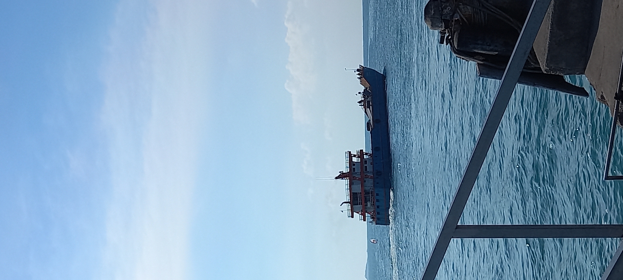 The image displays a clear maritime scene under a bright, expansive blue sky with a few faint, wispy clouds. In the mid-ground, a large blue and white cargo or supply ship is prominently featured, sailing across calm, rippled blue-green water. The vessel appears to be moving from right to left, evidenced by a discernible wake trailing behind it. A smaller, white boat, possibly a dinghy or tender, trails further behind the larger ship.
In the far distance, a subtle outline of land, likely hills or mountains, is visible on the horizon, hinting at a coastal or island setting. The foreground on the right side of the frame is occupied by parts of a dark, metallic structure, consisting of beams and railings. This suggests the photograph was taken from a pier, a dock, or the deck of another vessel, looking out over the sea.
The scene depicts a peaceful daytime setting, likely mid-morning or early afternoon, characterized by clear weather and good visibility. No specific human activity is distinctly visible on the ships, though the movement of the large vessel implies maritime operations. There is no visible text within the image. This tranquil view is consistent with coastal waters, such as those found near Thong Sala, Thailand.