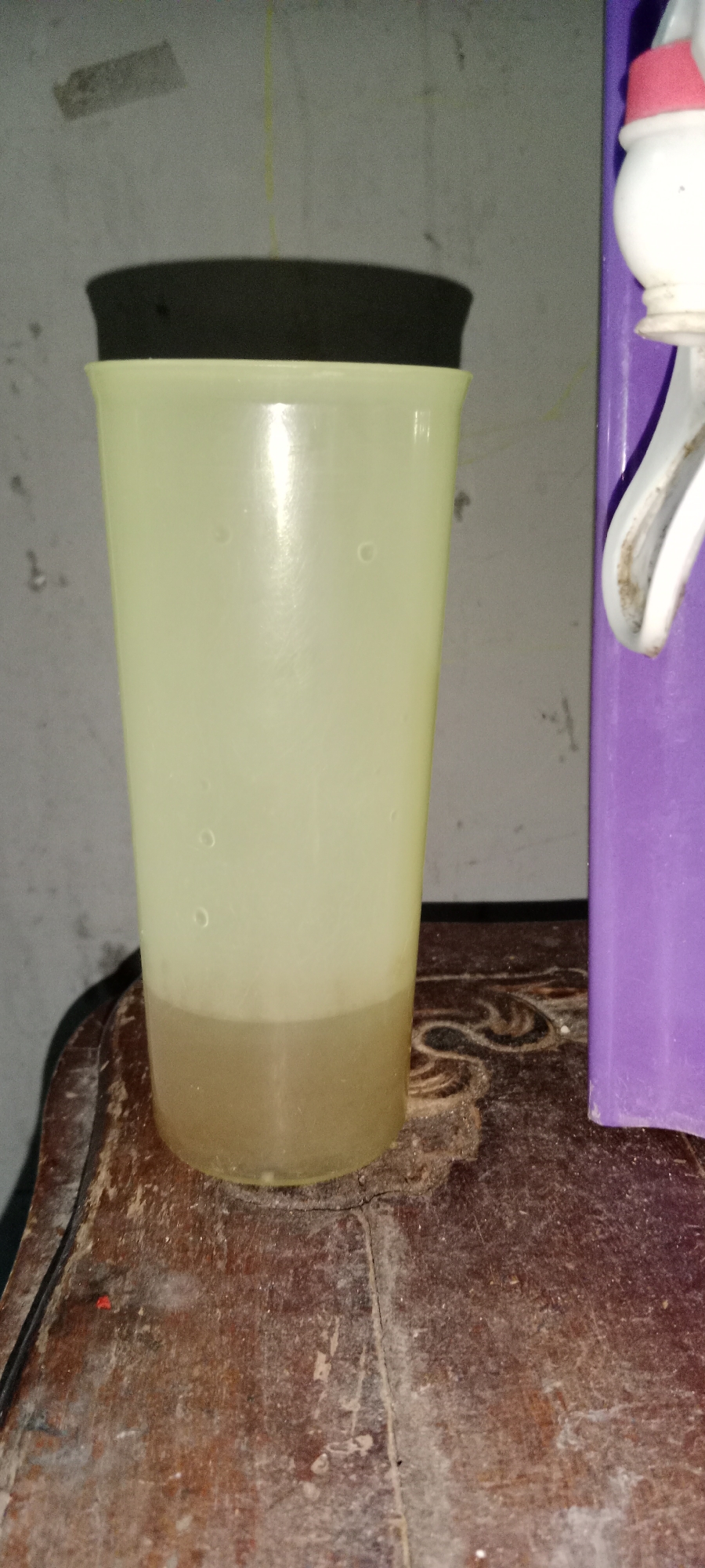 A tall, translucent yellow plastic cup with a black lid sits on a weathered wooden surface. The cup contains a light brown liquid, filling approximately the bottom third of the container. The wooden surface appears aged and possibly stained, with visible cracks and a layer of dust or debris. To the right of the cup, a portion of a purple appliance, likely a water dispenser, is visible. The background consists of a gray wall with some marks and a faint yellow line running vertically. The scene is indoors and appears to be in a residential or simple commercial setting. The lighting suggests it is either evening or the room is dimly lit. No people are present in the image.