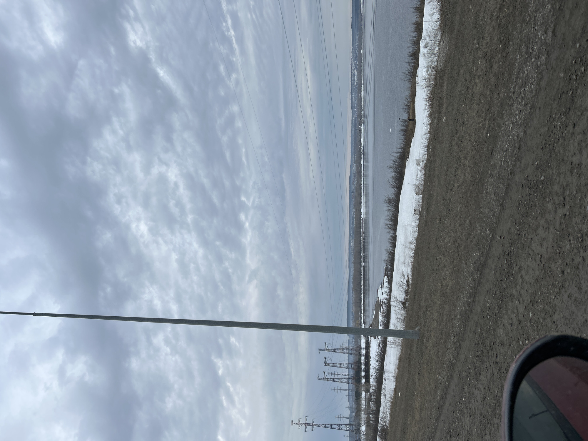The image presents a landscape view likely taken near the city of Chaikovsky, Russia, given the Volga River’s presence. The scene is dominated by an overcast sky filled with patchy clouds. A large body of water, likely the Volga River, stretches across the midground, bordered by a shoreline covered with patches of snow. A dirt or gravel road runs along the right side of the frame, and the rearview mirror of a vehicle is visible in the lower right corner. Power lines and utility poles are visible against the sky. The general ambiance suggests a cold, possibly late-winter or early-spring day. No people are visible.