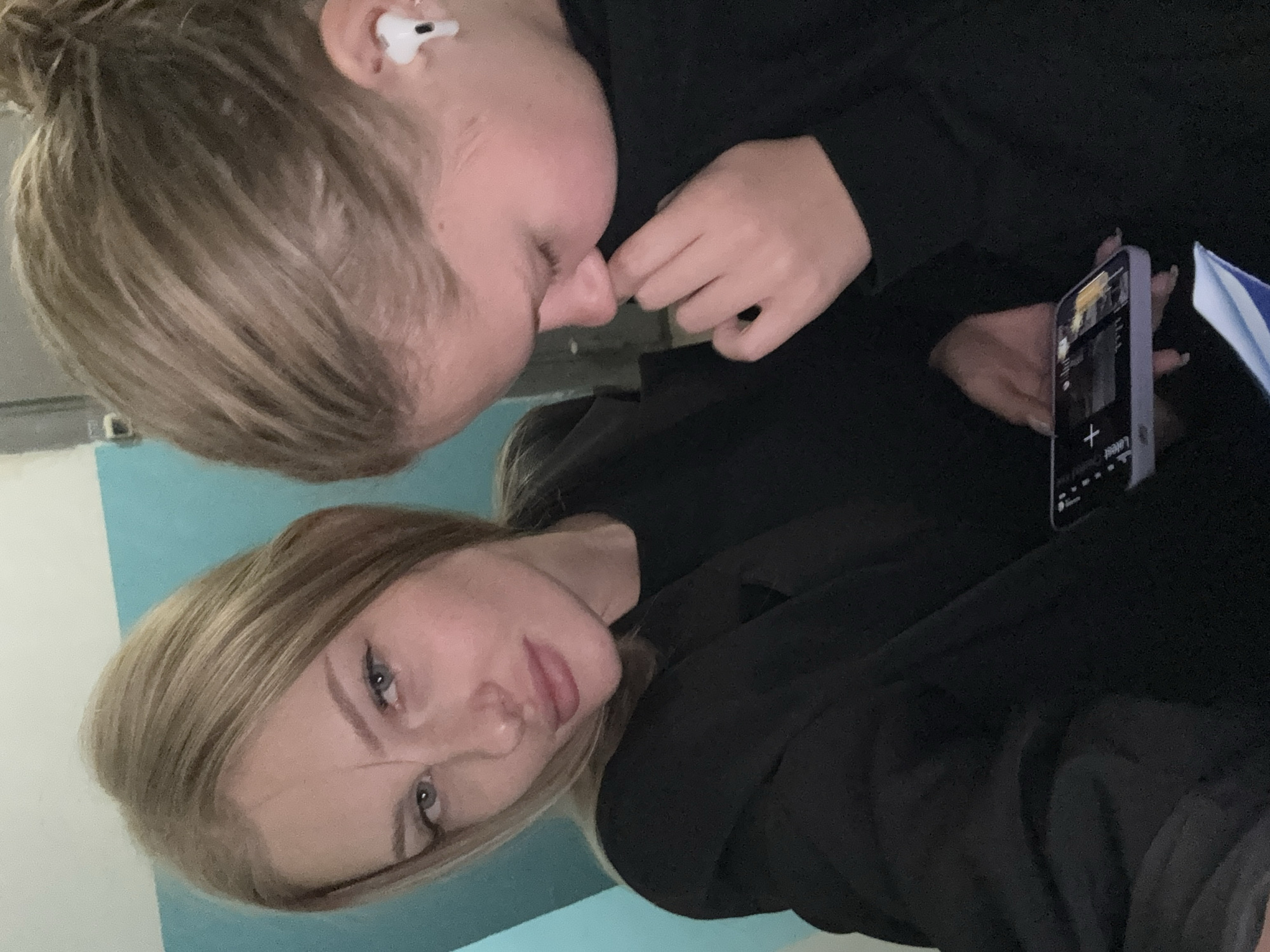 Two young women are captured in a selfie. The woman in the foreground, on the left, has blonde hair and is looking directly at the camera with a neutral expression. She is wearing a black jacket over a black top. The second woman, to the right, has her head bowed, and her face is partially obscured as she touches her nose. She is wearing a black top and a white wireless earbud in her right ear. She is holding a smartphone in her right hand, which displays a screen with various icons and text. The background consists of a wall with a turquoise section and a lighter section, suggesting an indoor setting, possibly a room or hallway. The lighting is artificial and appears to be coming from above. There are no distinctive landmarks visible to indicate a specific location in Perm, Russia.