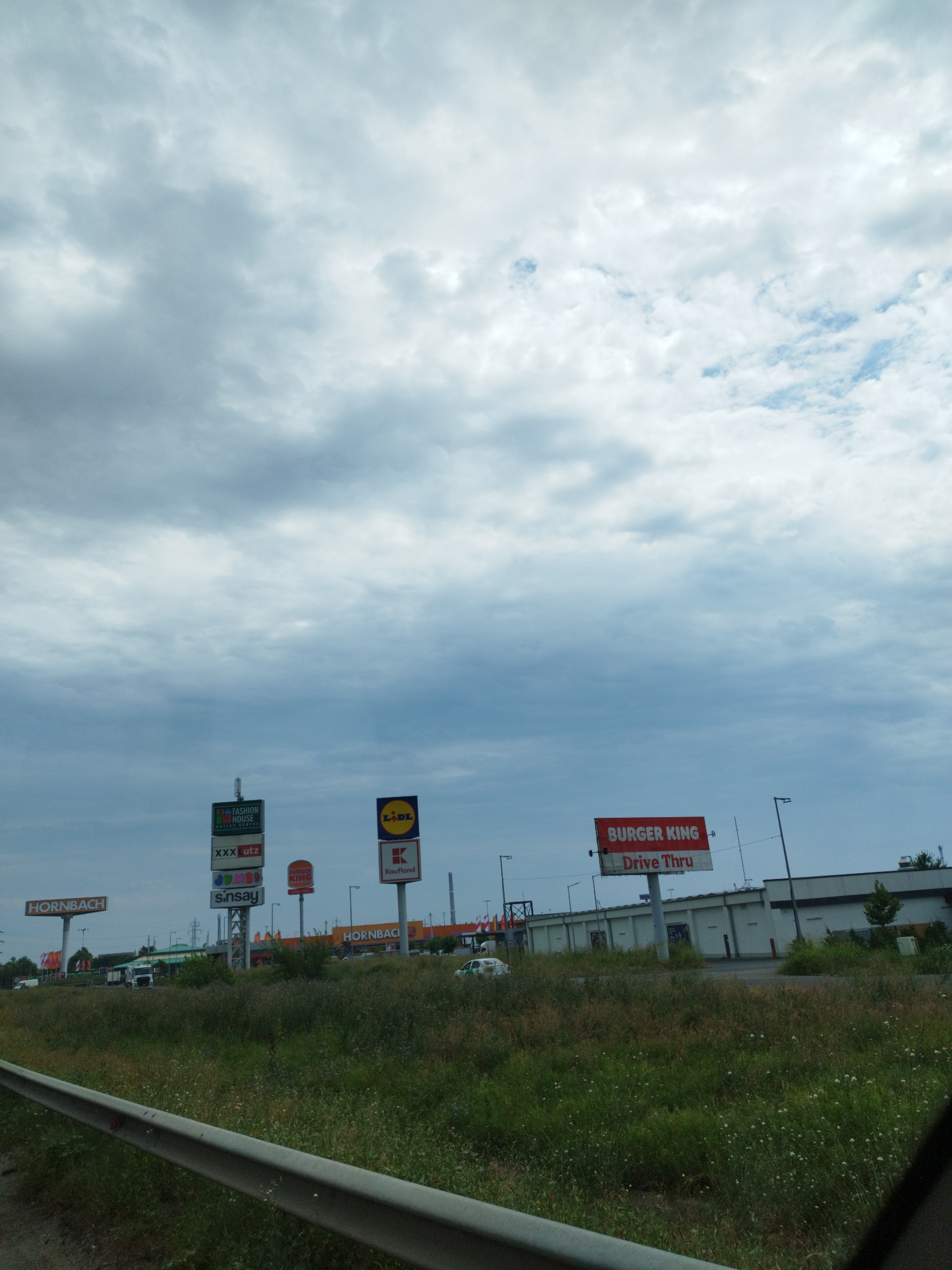 This is a photograph taken in Chiajna, Romania, depicting a commercial area under a cloudy sky. The foreground shows a grassy verge with a metal guardrail. In the midground, a row of commercial establishments is visible, characterized by various signage. Prominently displayed are signs for "HORNBACH" on the left and right. A cluster of vertical signs features "FASHION HOUSE", "XXX utz", and "Sinsay". Other visible signs include "Lidl" and "Kaufland". A "BURGER KING Drive Thru" sign is also present. Several vehicles, including trucks and cars, are parked and driving within the commercial complex. The background consists of buildings and more signs. The overall environment suggests a retail park or shopping center. There are no people clearly visible in the image.