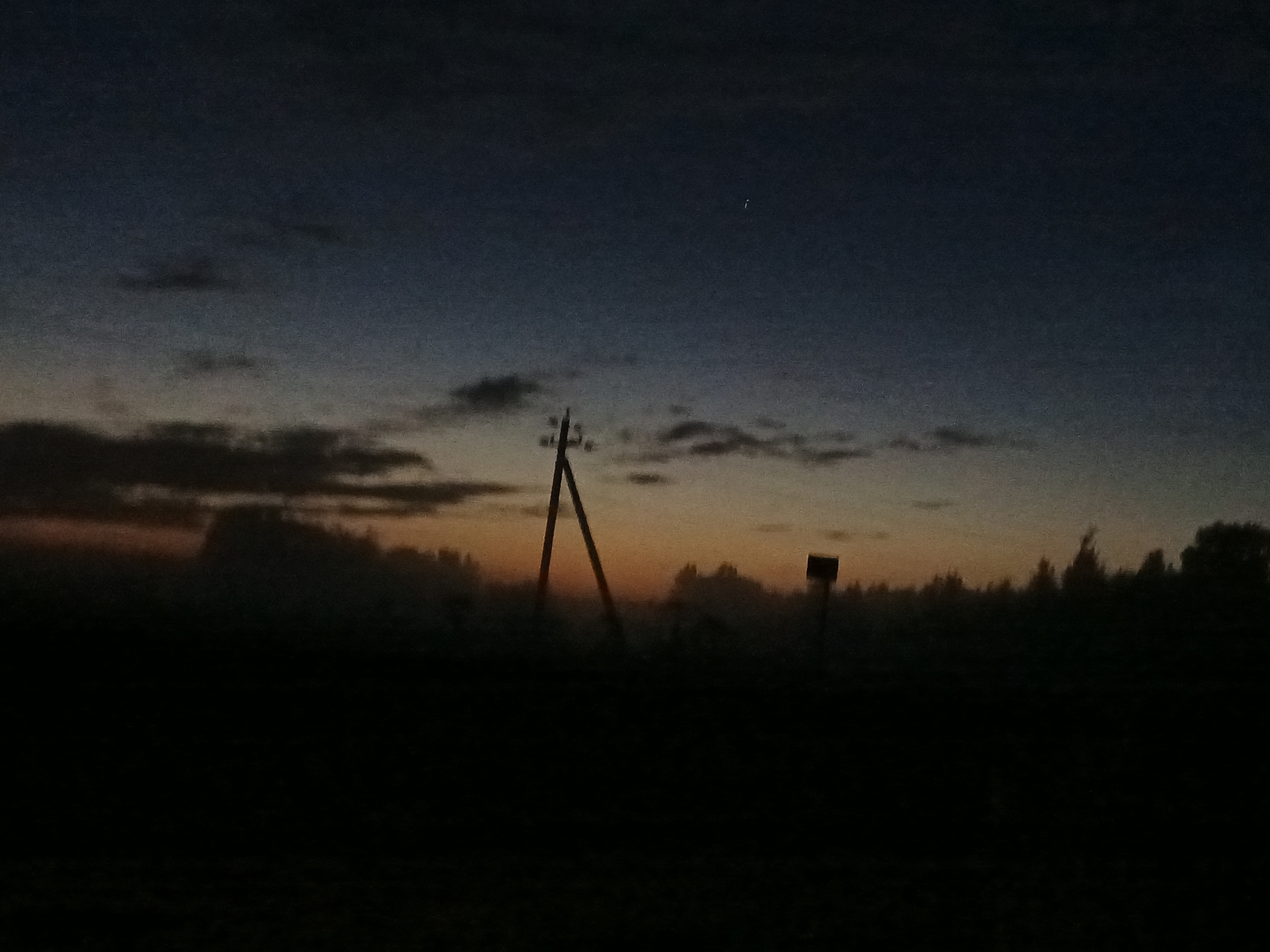 The image captures a dimly lit twilight scene in Воскресенское, Russia. The sky transitions from a dark blue overhead to a lighter orange and yellow near the horizon, indicating either dawn or dusk. Silhouetted against the sky are several elements: on the left, a tall, angular wooden structure, possibly a power line pole or a rustic signpost, stands prominently. To its right, further into the background, is a smaller, dark, square-shaped object mounted on a post, likely another sign. A line of dark trees forms a silhouette along the horizon, interspersed with what appears to be fog or mist obscuring the lower parts of the landscape. The foreground is almost entirely black, suggesting it is a field or unlit ground. No people or animals are visible in the frame. The overall atmosphere is one of quiet solitude.