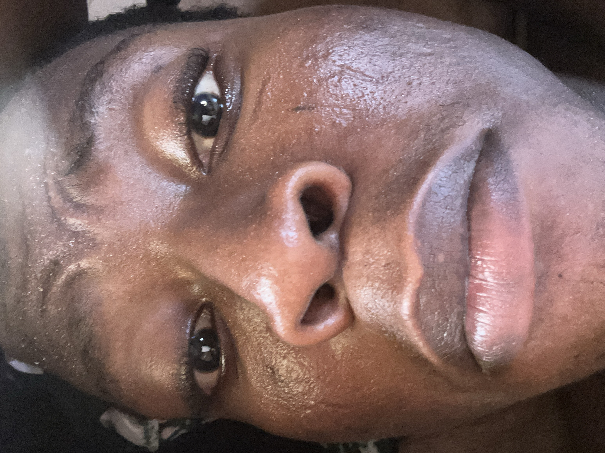 A close-up portrait of a person with dark skin, focusing on the face. The person has dark eyes, a wide nose, and full lips. The skin appears shiny and possibly sweaty. The person is looking upwards. The background is dark and out of focus, with some dark hair visible at the top. There is no indication of a city or location.