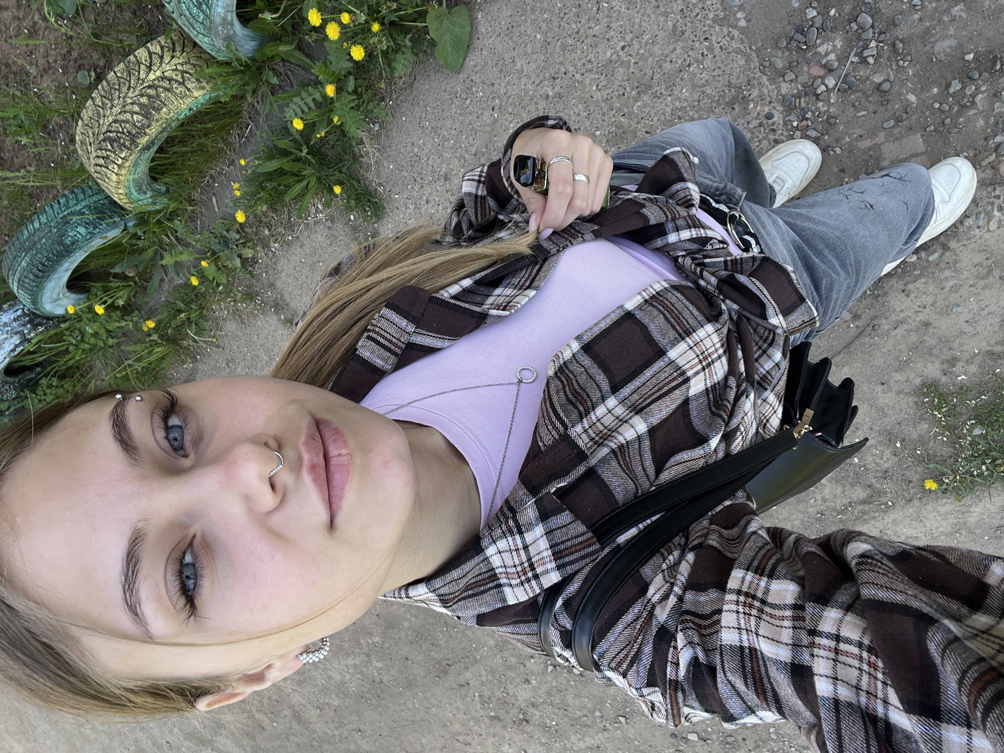 A young woman is posing in a selfie. She has fair skin, blonde hair, and blue eyes. She has several piercings in her face, including an eyebrow piercing, a nose ring, and earrings. She is wearing a light purple t-shirt and a brown and white plaid shirt, which is open. She is wearing gray jeans and white sneakers. She is also wearing several rings on her left hand. She is holding the collar of her shirt with her left hand. She has a black purse slung over her shoulder.
The background is a concrete surface, with some grass and yellow flowers in the upper left corner. There are some painted tires in the grass. The location cannot be determined from the image.