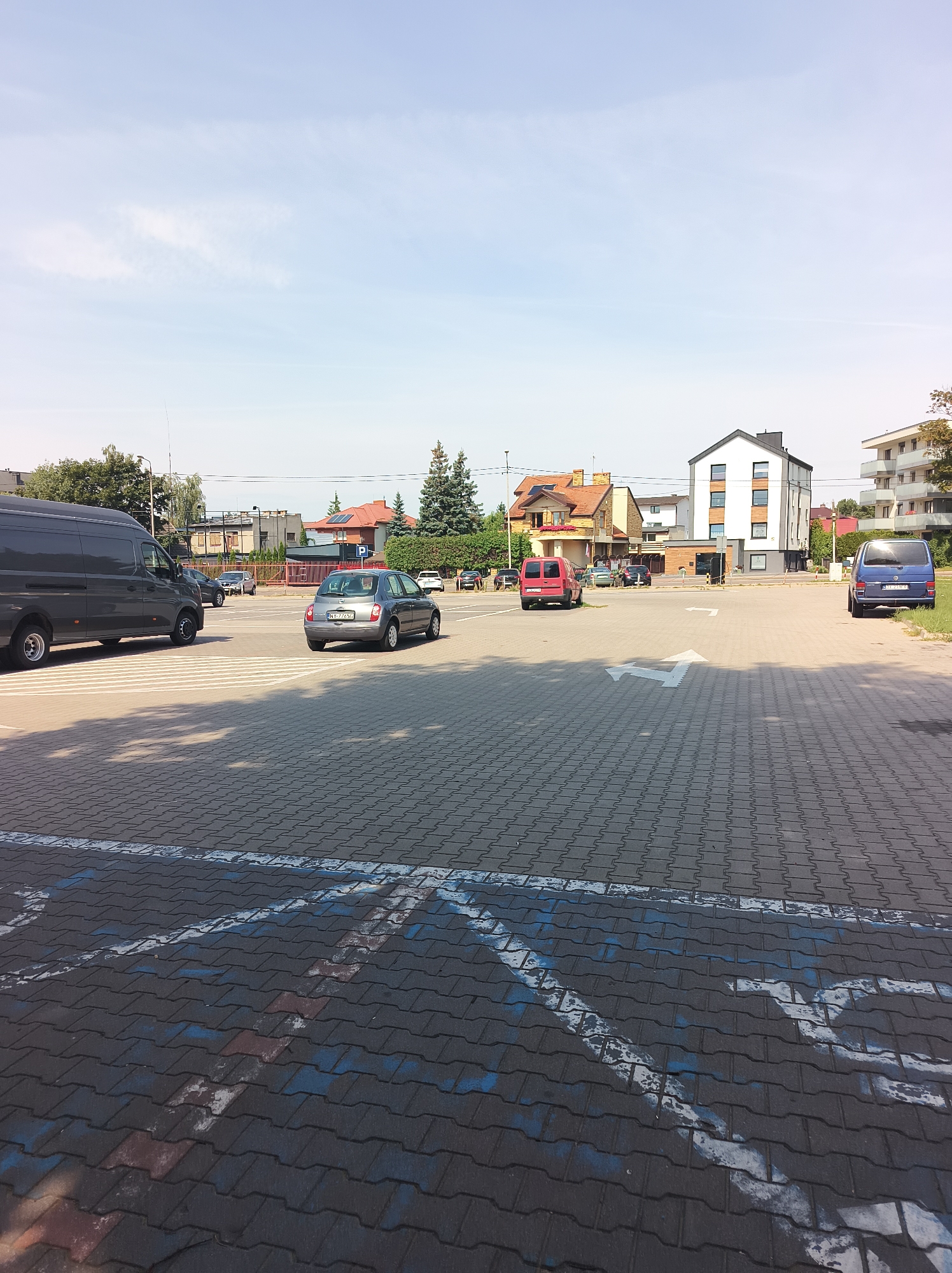 The image shows a parking lot with several vehicles and buildings in the background. In the foreground, the parking lot is paved with interlocking concrete pavers. White and blue markings are visible on the ground, indicating parking spaces.
On the left side of the image, a dark gray panel van is parked. To the right of the van, a gray hatchback car is driving away from the camera. Further to the right, a red panel van is parked. In the distance, a blue panel van is parked on the right edge of the frame.
Several buildings are visible in the background. On the left, there are houses with red-tiled roofs and trees. In the center, a modern, multi-story white building with dark windows stands out. To its right, another multi-story building with balconies is partially visible. The sky above is light blue with some wispy clouds. The overall scene appears to be a commercial or residential area on a sunny day.
The image is identified as being taken in Białystok, Poland, specifically on Storczykowa Street. This area features a mix of residential and commercial structures. The parking lot itself appears to be adjacent to buildings, possibly serving a residential complex or a business. The vehicles present are common types seen in urban environments. The presence of what appear to be disabled parking symbols (blue and white markings) suggests a public parking area.