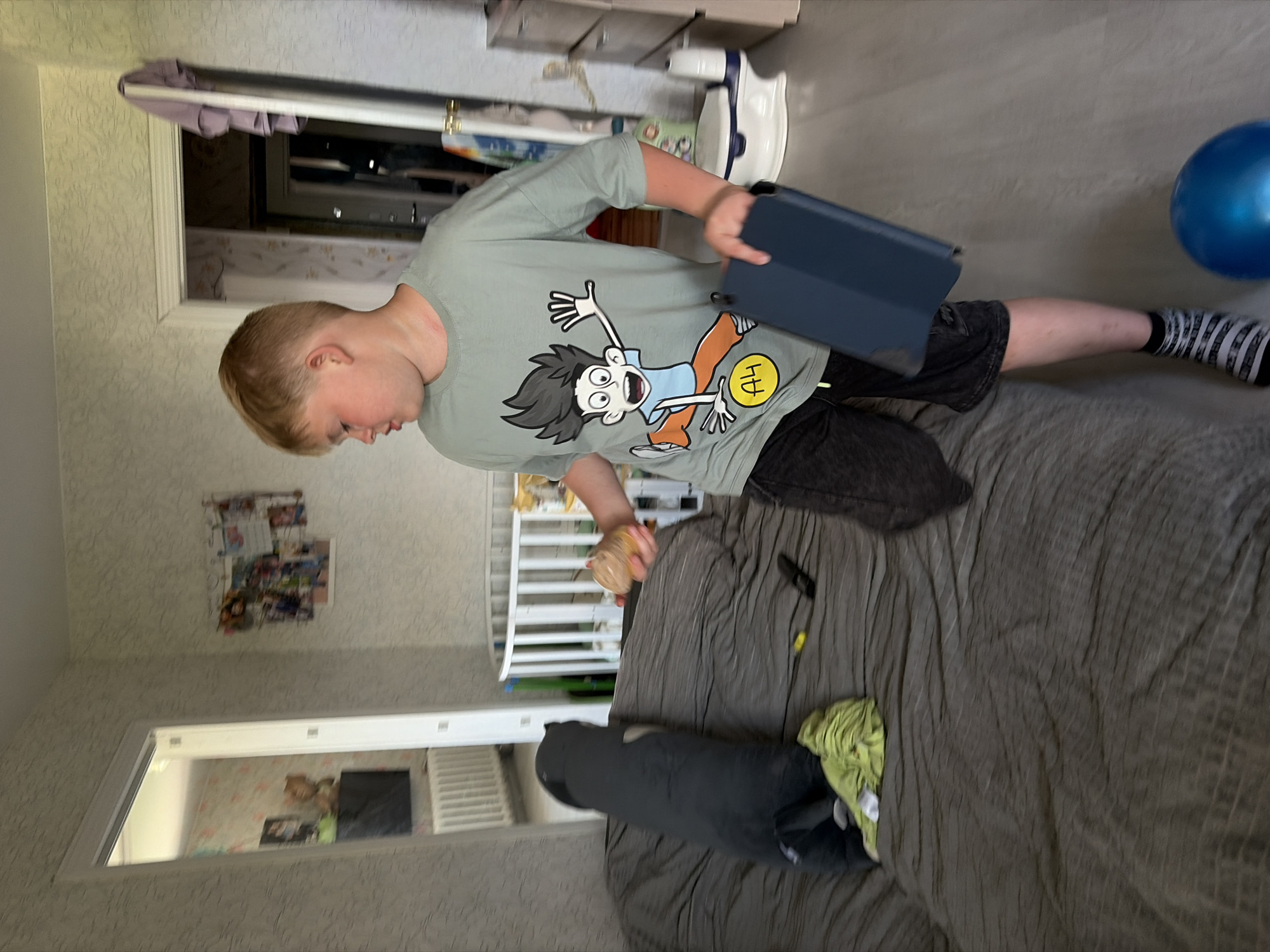 A young boy with blonde hair and a light green t-shirt with a cartoon character print is standing on a wooden floor in what appears to be a bedroom or play area. He is holding a dark blue tablet in his right hand and a light brown object, possibly an ice cream cone, in his left hand. He is wearing dark shorts and striped socks. In the background, there is a bed with a dark gray comforter, a long dark gray pillow, and a green cloth. A white crib is visible further back, along with a door leading to another room. A blue balloon is on the floor near the boy's feet. The room is decorated with wallpaper and framed pictures on the wall.