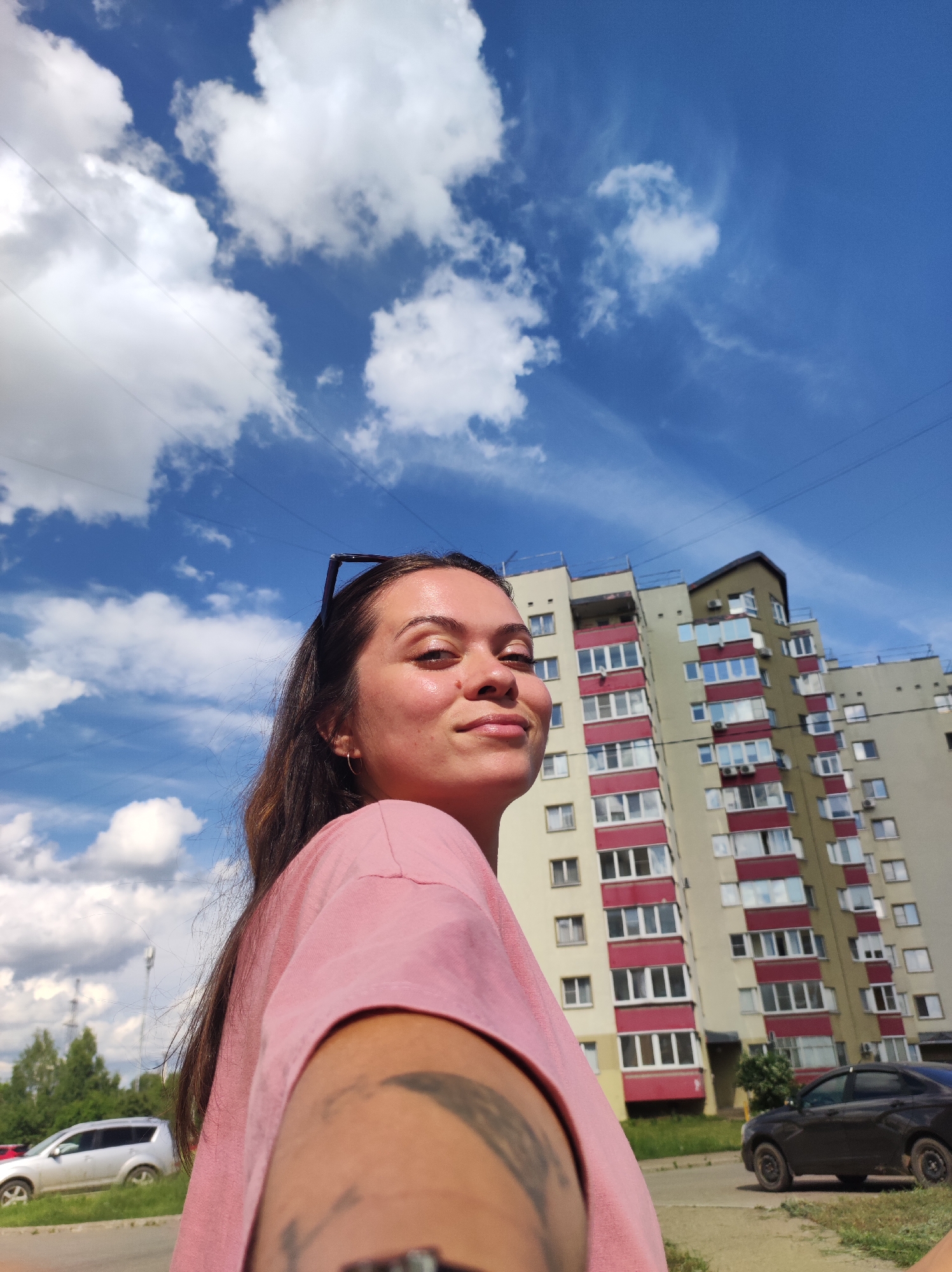 A young woman with long, dark hair is taking a selfie outdoors in the city of Chaikovsky, Russia. She is wearing a pink t-shirt and has sunglasses perched on her head. Her left arm, with a tattoo visible, is extended towards the camera. Behind her, a multi-story apartment building with yellow and red accents dominates the background. The sky is bright blue with scattered white clouds. A dark-colored car is parked on the street in front of the building, and a silver SUV is visible further down the road, alongside trees and a utility pole. The scene appears to be a sunny day.