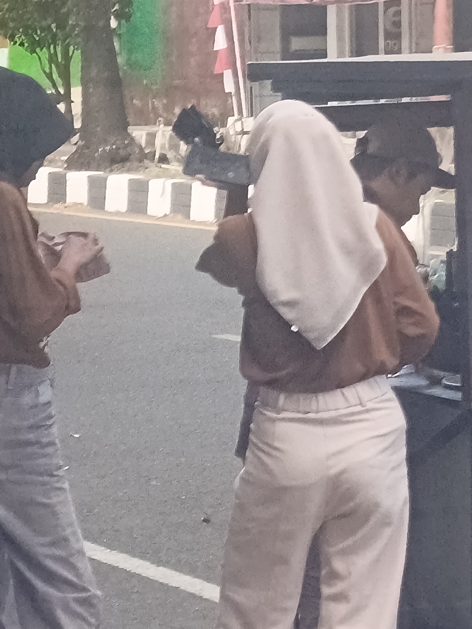 Two individuals are visible in an outdoor urban setting, likely in Desa Gunungsimping, Indonesia. One person, on the left, wears a dark headscarf and a brown shirt, and is holding a small reddish object in their hands. They are positioned next to a grey sidewalk with white rectangular dividers. The second person, on the right, wears a light-colored headscarf, a brown shirt, and light-colored pants. They are standing near a dark structure, possibly a food cart or stall, and appear to be interacting with something on or within it. A tree is present in the background, as well as a building with white and red elements, possibly flags or banners. The ground is paved and appears to be a road or walkway.