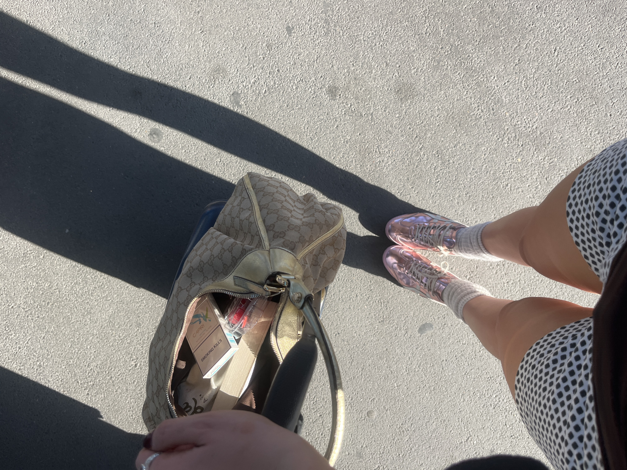 The image is a high-angle shot taken outdoors on a sunny day. The location appears to be a city street with a light gray asphalt surface. Two dark shadows stretch diagonally across the left side of the frame, suggesting the presence of nearby structures or objects casting the shadows.
A person's legs and feet are visible on the right side of the frame, wearing a white and black patterned skirt, white socks, and pinkish-purple sneakers.
To the left of the person's legs is a partially open beige Gucci handbag. The contents of the bag are visible, including a pack of Capri cigarettes, some makeup items, and other miscellaneous objects.
The image does not contain any contextual clues indicating the city of Paris.