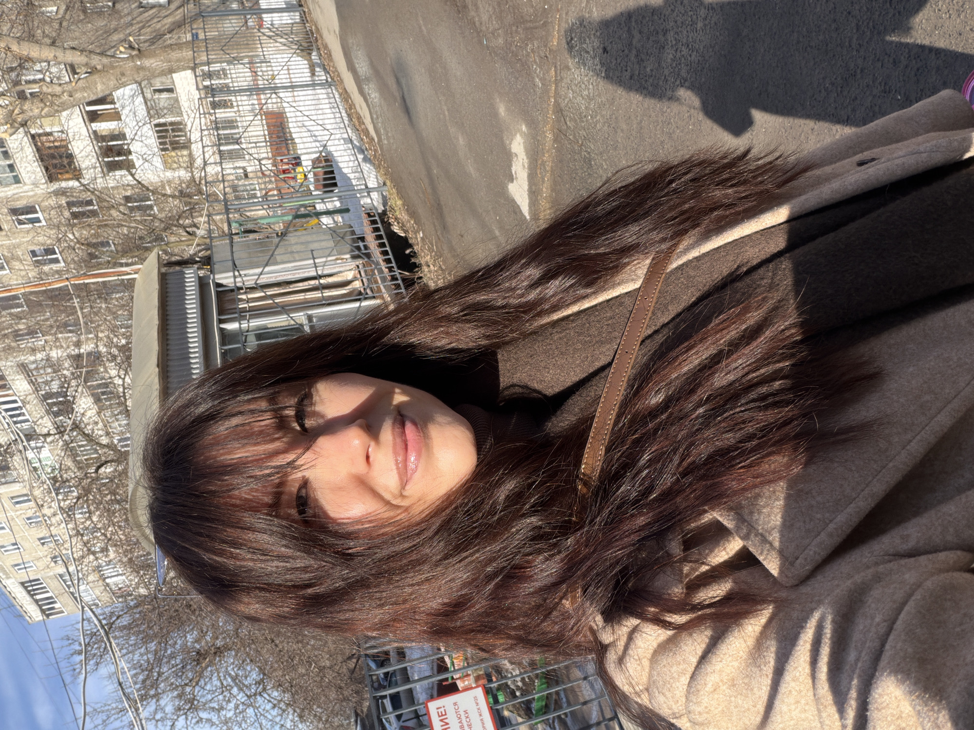 A female individual, likely in her late 20s to mid-30s, is captured in a selfie-style portrait on a clear, sunny day in Perm, Russia. She has long, dark brown hair with bangs, parted centrally, and is smiling directly towards the camera. She wears a light beige overcoat and a dark brown turtleneck or sweater, with a brown leather strap visible over her right shoulder. Direct sunlight illuminates her face from the right, casting defined shadows.
The background depicts an urban residential environment. A multi-story, light-colored apartment building, consistent with Soviet-era architecture, stands prominently. Bare deciduous trees are scattered throughout the scene, and patches of snow are visible on the ground, indicating a colder season. A metal fence runs horizontally across the mid-ground, behind which lies a children's playground with glimpses of red and yellow equipment and a small, corrugated metal utility structure. An asphalt surface, showing signs of wear and minor debris, forms the foreground. Overhead, numerous utility wires crisscross against a bright blue sky. A partial red and white sign with Cyrillic text, including "ЖСК №25," is visible on the far left, indicative of a Russian housing cooperative. The photographer's shadow is distinctly cast on the asphalt surface to the lower right.