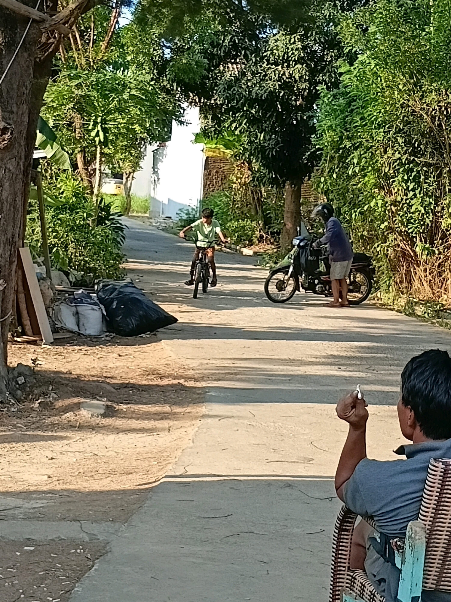 In Cikal, Indonesia, a young person on a bicycle rides down a paved road bordered by lush green trees. To the right, a person wearing a helmet and a blue shirt stands next to a motorcycle. In the foreground, a man sits in a wicker chair, smoking a cigarette, with trash bags and discarded materials visible to the left of the road. The scene is bathed in the warm sunlight of what appears to be late afternoon.