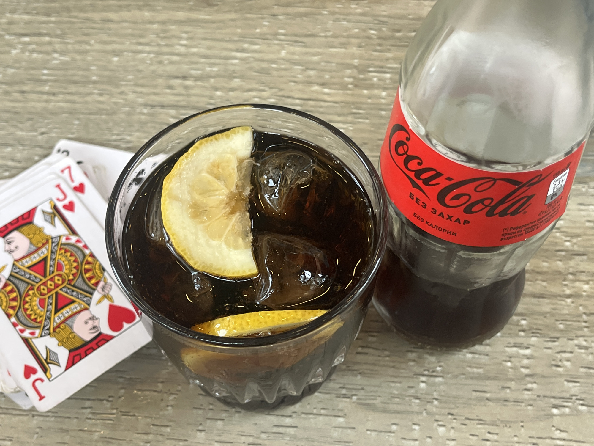 An overhead shot captures a glass of Coca-Cola with ice and lemon slices, a Coca-Cola bottle, and a stack of playing cards on a light-colored wooden surface.
The glass, positioned on the left side of the frame, contains a dark brown beverage, presumably Coca-Cola. Several ice cubes are visible, along with two slices of fresh lemon, one resting on the rim of the glass and another submerged. The glass itself has a textured, faceted design on its lower half.
To the right of the glass stands an empty, clear glass Coca-Cola bottle. Its red label is prominent, featuring the "Coca-Cola" logo in white script. Below the main logo, text in Cyrillic script reads "БЕЗ ЗАХАР" (SUGAR FREE) and "БЕЗ КАЛОРИИ" (NO CALORIES), indicating it's a "Coca-Cola Zero" product. Further smaller Cyrillic text, partially visible, includes words like "Енергия" (Energy), "%0", "Референтни" (Reference), "прием на" (intake of), and "възрастен" (adult), likely detailing nutritional information or recommended daily intake.
To the left of the glass, a stack of playing cards is partially fanned out. The topmost card is clearly visible, displaying the "Jack of Hearts" (J♥). Below it, another card shows the "7 of Hearts" (7♥). The cards are standard design with red and black motifs.
The setting appears to be indoors, with the objects arranged on a light brown, wood-grained table or countertop. The lighting is bright and even, suggesting an indoor environment. The image suggests a casual activity, perhaps a break or a game of cards accompanied by a refreshing drink.
