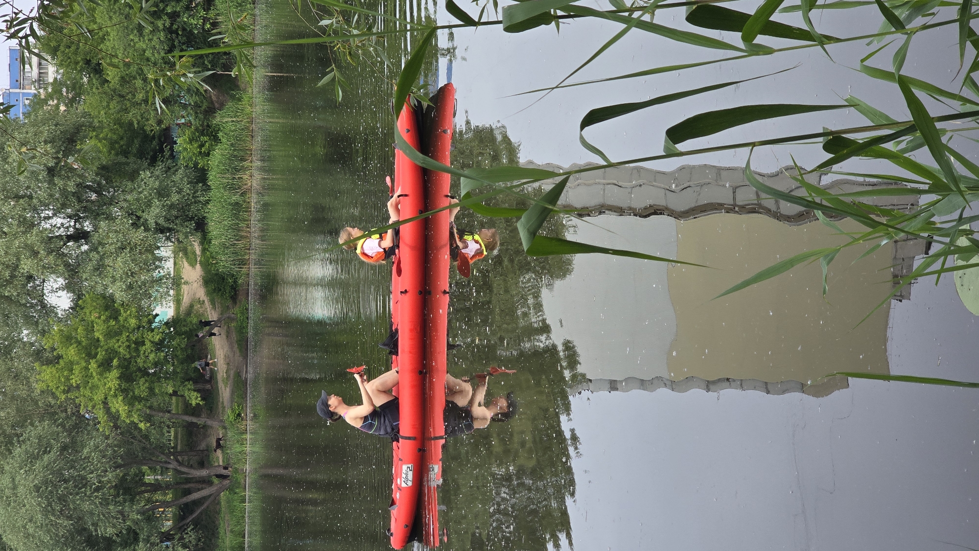 The image captures two individuals in a red inflatable kayak on a body of water, likely the Rusanivka Channel, in Kyiv, Ukraine. An adult female, wearing a dark cap, dark athletic top with colorful straps, and shorts, is seated in the front-left position, holding a red object. A child, wearing a light-colored life vest and a light top, is seated in the front-right position. Both individuals are looking generally towards the right side of the frame. The brand "Alpha" is visible on the side of the red kayak.
The water is dark and exhibits slight ripples, reflecting the kayak, the individuals, and the surrounding environment. The far bank is covered with dense green vegetation, including large deciduous trees with light green and silvery foliage, and a thick band of reeds along the waterline. A dirt path is visible on this bank, with several indistinct figures and a dog present in the distance on the top-left.
In the background, behind the trees, multi-story urban residential buildings are visible, including a light blue/grey apartment building on the upper right. The foreground on the right side of the image features blurred green reeds and grass blades, partially obstructing the view of the water and reflections. A prominent, distorted, light-colored, multi-story building with vertical structural elements is reflected distinctly in the lower half of the water, a common sight along the Rusanivka Channel's urbanized banks. The sky appears overcast.