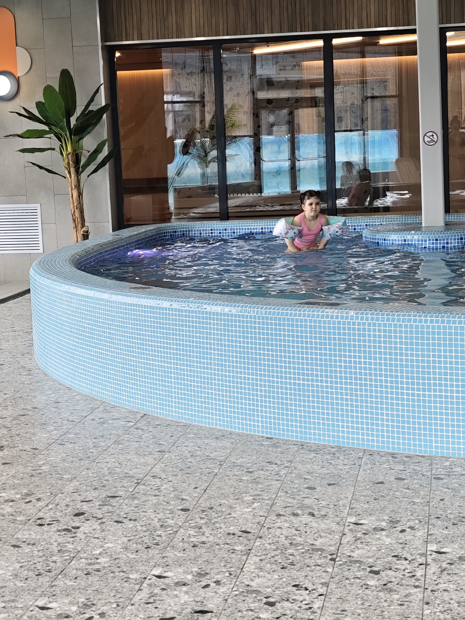 The image captures a young girl in a blue-tiled, curved indoor pool, likely located in Votkinsk, Russia. The girl, appearing to be a toddler, wears a pink swimsuit and white and blue arm floats. She is positioned in the center of the frame, partially submerged in the rippling water, looking directly at the camera with a calm expression.
The pool itself is circular or semi-circular, lined with small, light blue mosaic tiles. The surrounding floor is covered in large, light grey, speckled tiles. In the background, a modern interior facility is visible. To the left, a large potted plant with broad green leaves stands next to a grey tiled wall with a white ventilation grate. Above, an orange and grey circular decorative element is affixed to the wall.
Further in the background, large glass panels or windows reflect various elements, including what appears to be another section of the facility with wooden paneling and indistinct shapes. Reflections of a bright blue color, possibly from the sky or an outdoor pool, are also visible through the glass. A white vertical pillar on the right features a circular "no diving" or "no jumping" sign. The ceiling above the glass is adorned with light brown wooden slats. The lighting suggests an indoor daytime setting, with a bright ambiance.