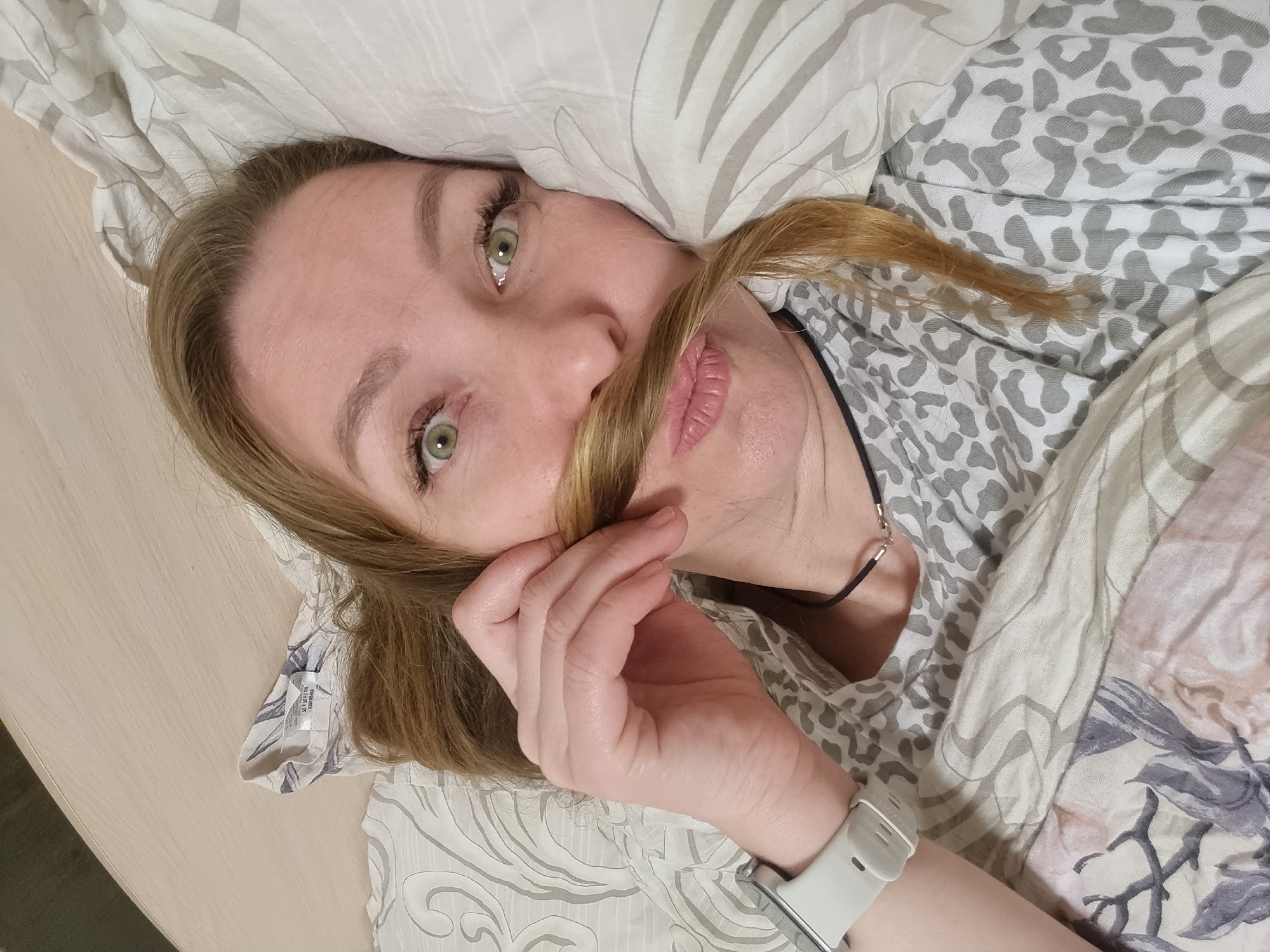 A woman with light brown hair and prominent green eyes lies down, presumably in a bed, looking directly at the camera. She has a playful expression, holding a strand of her hair across her upper lip with her right hand, mimicking a mustache. Her lips are softly pink. She wears a thin, dark necklace with a small golden clasp and a light grey smartwatch on her left wrist.
Her head rests on a pillow with a light grey and white patterned pillowcase. She is partially covered by bedding that features a light grey leopard print on one section and a floral/abstract pattern in light pink, grey, and white on another. A light-colored wooden or laminate surface is visible to her left. The indoor setting suggests a bedroom, with soft, even lighting. No specific location cues for Chaikovsky, Russia are visible within the image, nor is any legible text apparent.