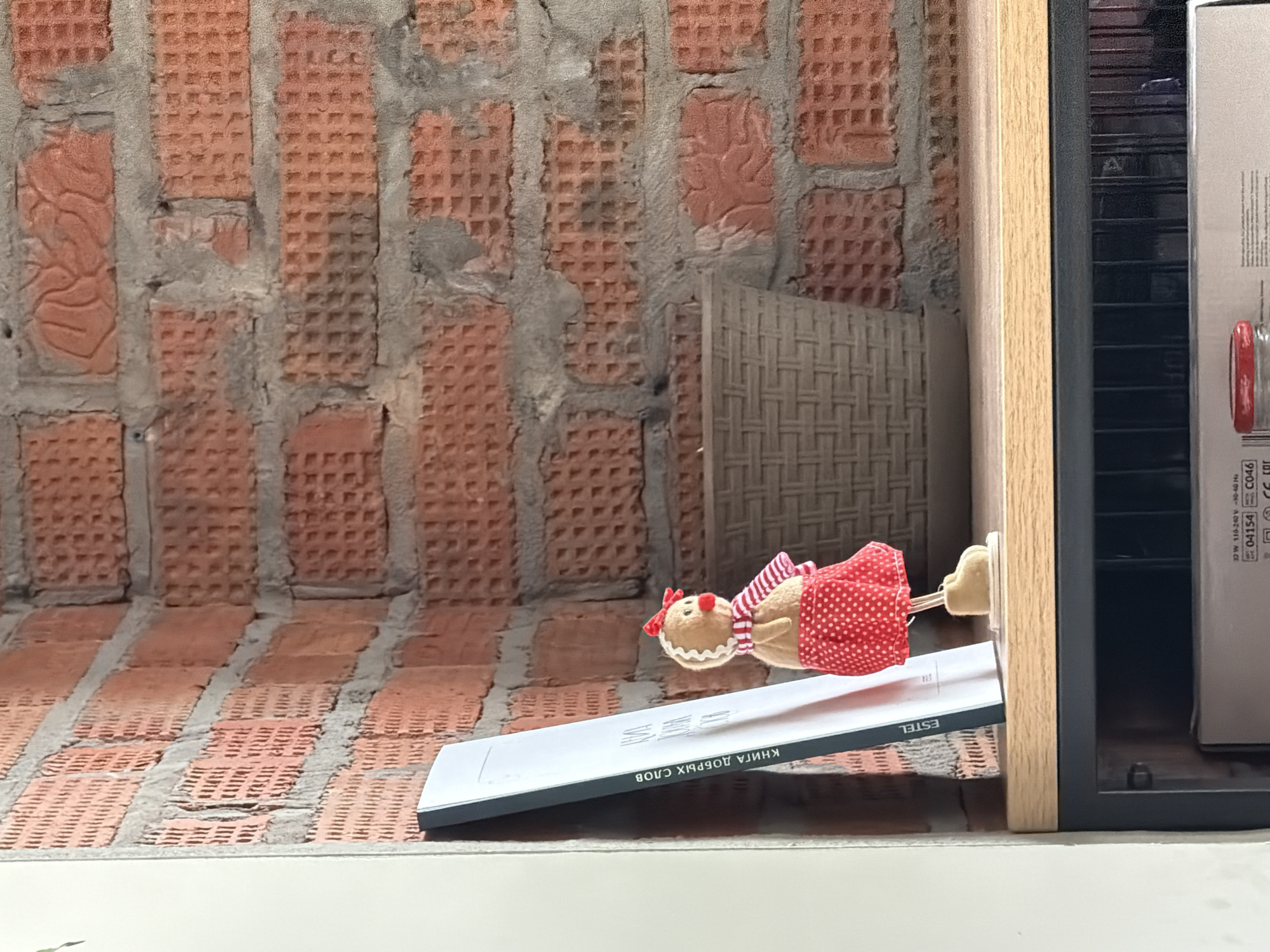 A gingerbread person doll sits on a white rectangular object with text in Russian that translates to "Book of Good Words" resting on a shelf within a wooden cabinet in front of a red brick wall. The setting is an interior space, seemingly in Pervomaysky, Russia.