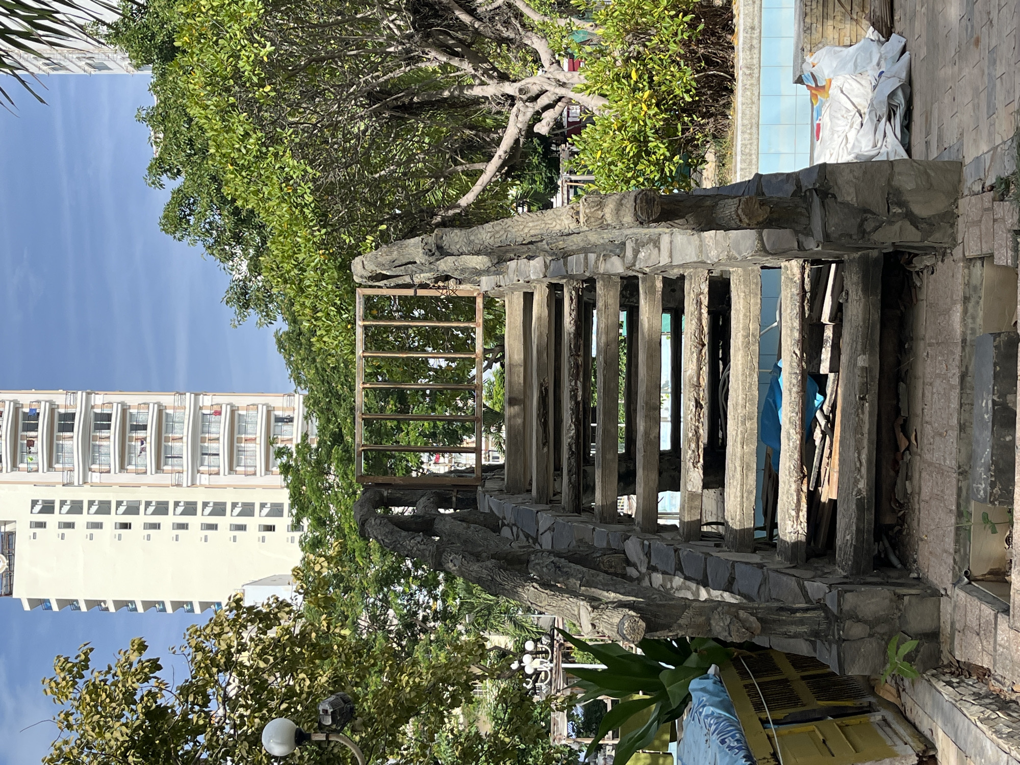 This image depicts an outdoor scene in Nha Trang, Vietnam, featuring a weathered stone staircase with ornate, textured railings resembling tree branches. The staircase leads upwards towards a rusty metal gate and is flanked by lush green trees and foliage. In the background, a tall, multi-story building with numerous balconies is visible, alongside a clear blue sky. The foreground shows the base of the stairs with debris and a partially visible swimming pool area.