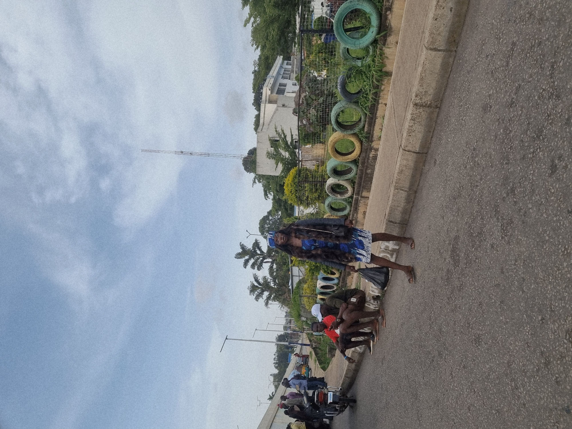 The media file depicts an outdoor scene in Jos, Nigeria, likely during the day, under a cloudy sky. The foreground is dominated by a paved road, bordered by a concrete curb and a grassy area. In the grassy area, a line of painted tires serves as a decorative or functional barrier.
Several people are present in the image. A woman, dressed in a blue and white patterned dress and a dark jacket, stands on the roadside, holding a black plastic bag. She appears to be wearing a headpiece. She is standing near a group of men who are seated or squatting along the curb. One man in the group is wearing a white cap and another is wearing a red shirt. Further down the road, a collection of motorcycles and people are visible, suggesting a transportation hub or a busy street. Some individuals are seated on the motorcycles, while others stand nearby.
In the background, there is a building with multiple windows and a telecommunications tower. Palm trees and other vegetation are also visible, contributing to the tropical or sub-tropical environment. The overall impression is one of everyday life in an urban or semi-urban setting in Nigeria, with people engaged in various activities like waiting, socializing, or preparing for travel. There is no visible text in the image.
