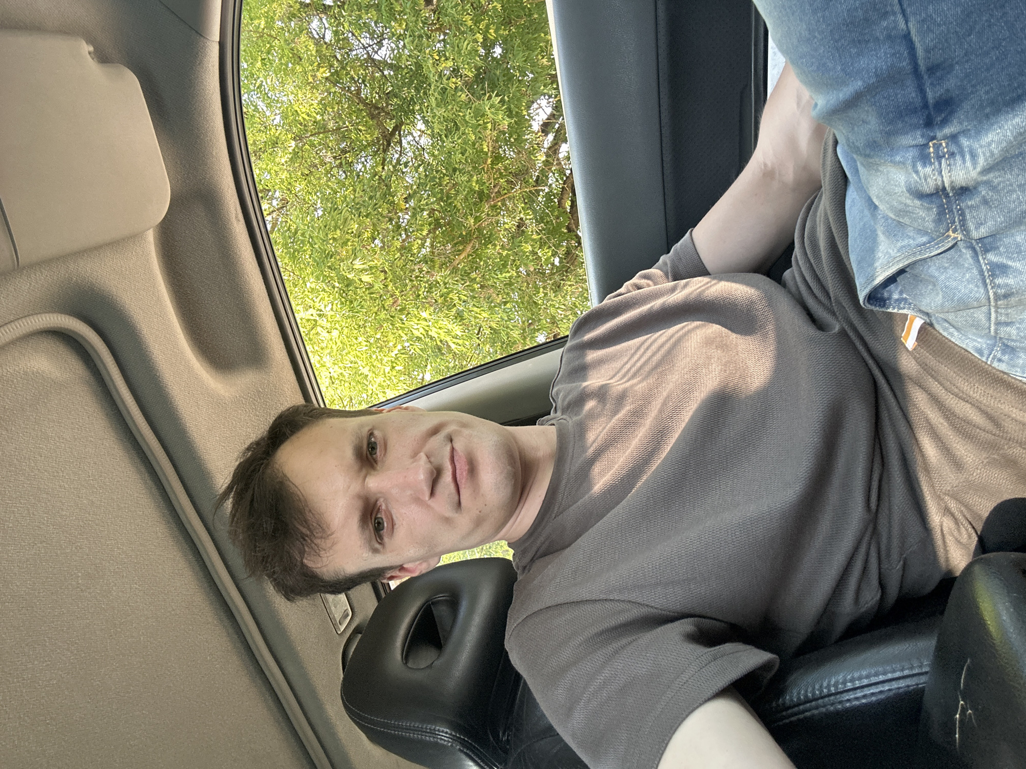 A man is sitting in the driver's seat of a car, looking at the camera. He has short brown hair and is wearing a grey t-shirt and blue jeans. The car's interior is visible, including the dashboard, steering wheel, and a black leather seat. Outside the car, a tree with green leaves is visible through the window. The image appears to be taken during the daytime, with natural light illuminating the scene. The specific location in the city of Makiv, Ukraine is not evident from the image itself, as there are no distinguishing landmarks or identifying signs.