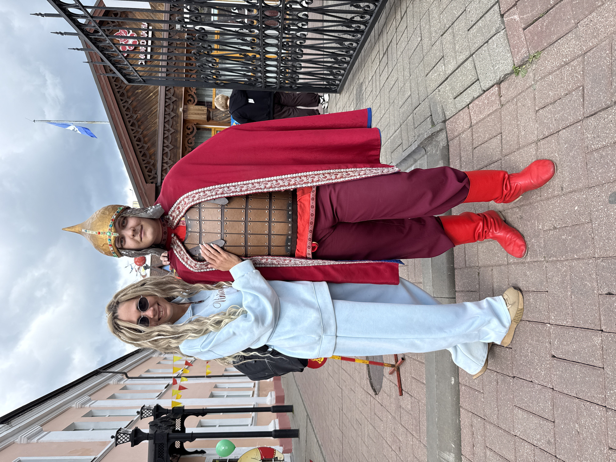 The image shows two individuals posing outdoors on a brick-paved surface. One person is dressed in historical attire resembling a medieval warrior, featuring a red cape with ornate trim, a helmet, and a leather-like breastplate. They also wear red boots and maroon-colored pants. The second person is dressed in a light blue tracksuit and sunglasses, with long blonde hair. They are standing next to the person in historical attire, with one arm around them.
Behind the subjects, there are architectural elements of a building, including a wrought-iron fence and a wooden structure with decorative details. Further in the background, there's a building with flags and colorful banners. The sky appears overcast.
Based on the available information, the location is Rusanovo, Russia. The overall scene suggests a tourist or cultural event.