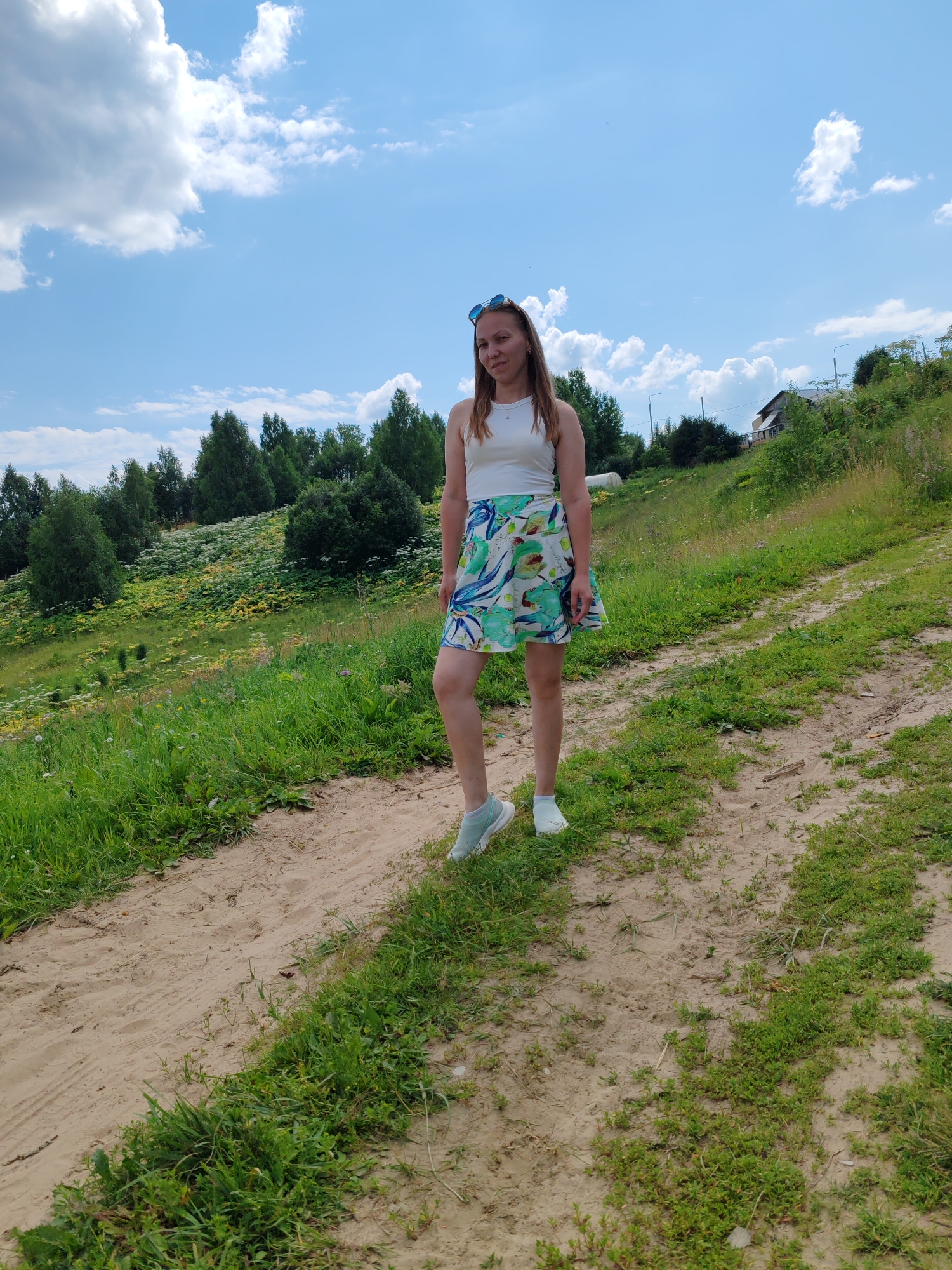 A woman stands on a dirt path on a grassy hill under a blue sky with scattered clouds. She is wearing a white tank top, a floral skirt with green, blue, and white patterns, and light blue sneakers with white socks. She has blonde hair and is wearing sunglasses on top of her head. The path is sandy and appears to be a dirt road or trail. The hill is covered in green grass and various wildflowers, including large white blooms, possibly Queen Anne's Lace, and yellow flowers. In the background, there is a line of green trees and some buildings on a higher part of the hill. The overall setting suggests a rural or natural environment, possibly on the outskirts of a settlement.