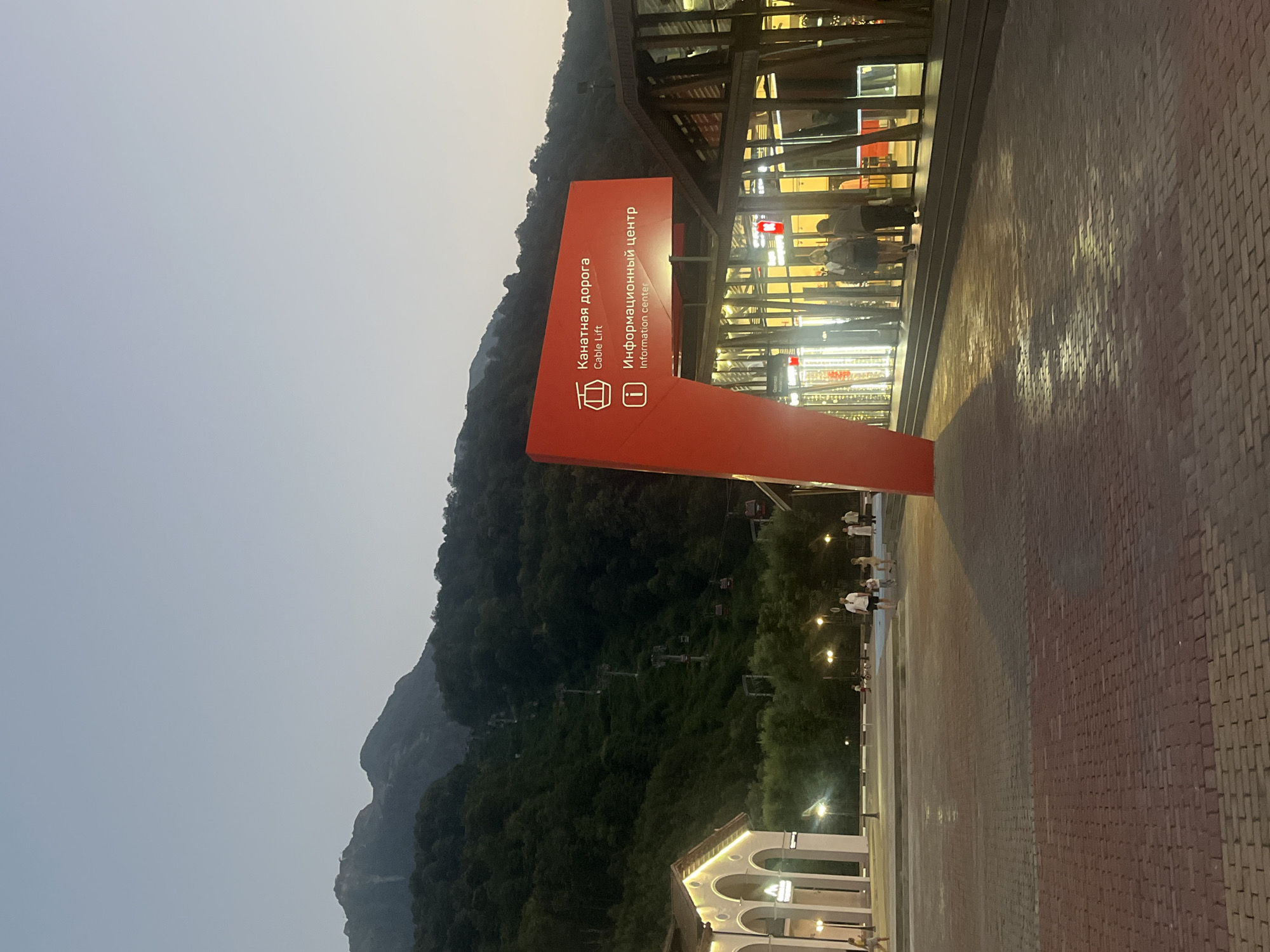 The image shows a cable car station in Estosadok, Russia. The station is located at the base of a mountain, and the cable cars can be seen ascending the mountainside. A large red sign at the entrance to the station displays information in Russian and English about the cable car and an information center. Several people are walking around the plaza in front of the station, and the area is lit by streetlights. The sky is dusk, and the mountains are covered in trees. The overall scene depicts a busy tourist destination.