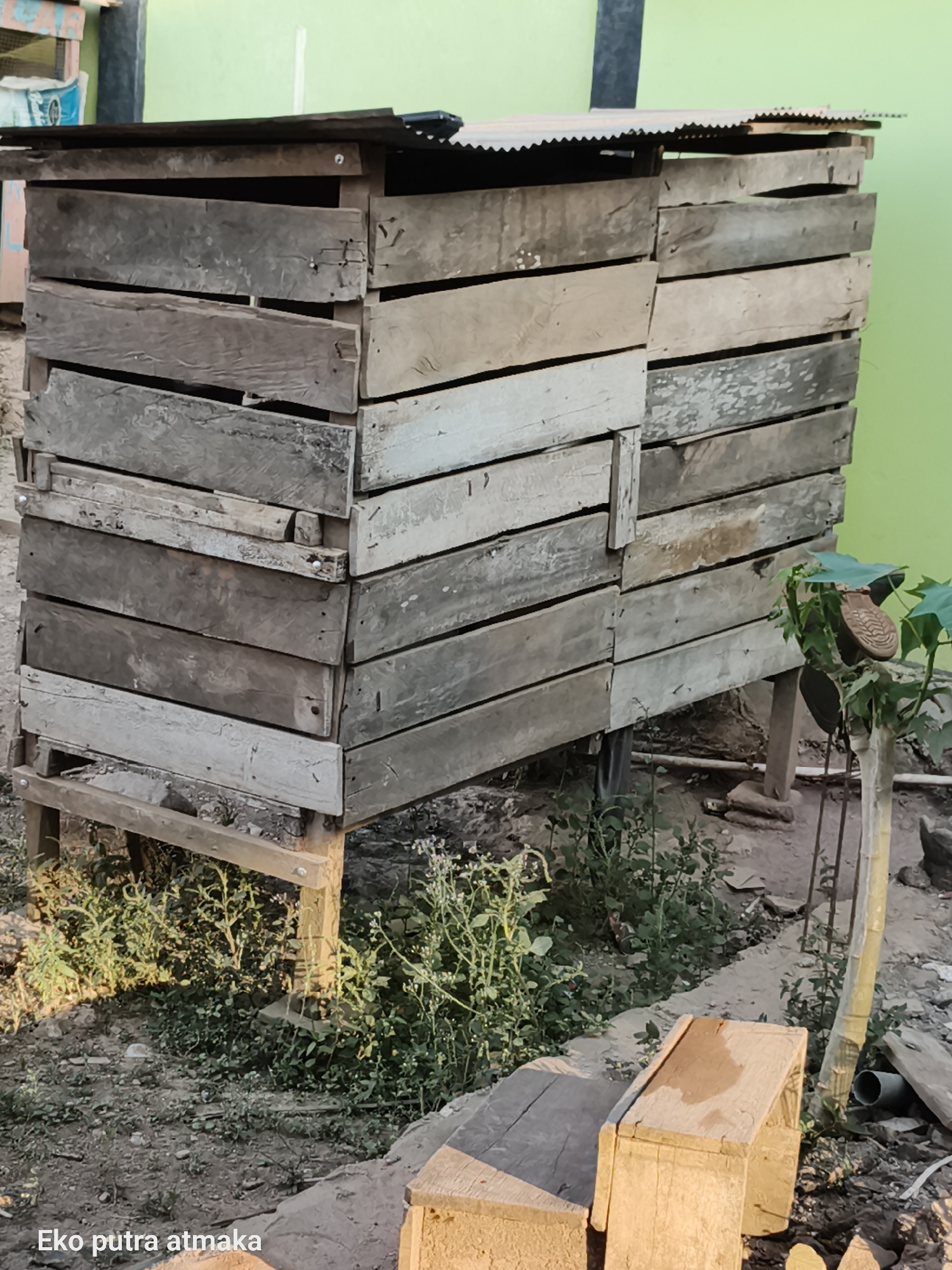This is a medium shot, eye-level, outdoor photograph taken in Pelanjau Jaya, Indonesia. The main subject is a rustic, multi-tiered structure made of weathered wooden planks, likely serving as a raised coop or storage unit. The structure is supported by four wooden legs and has a corrugated metal roof. It is surrounded by dry soil, scattered vegetation, and some building materials. To the right of the structure, a small plant grows with a shoe sole hanging from one of its branches. The background features a light green wall. In the foreground, a wooden object is partially visible. The scene appears to be during daylight.