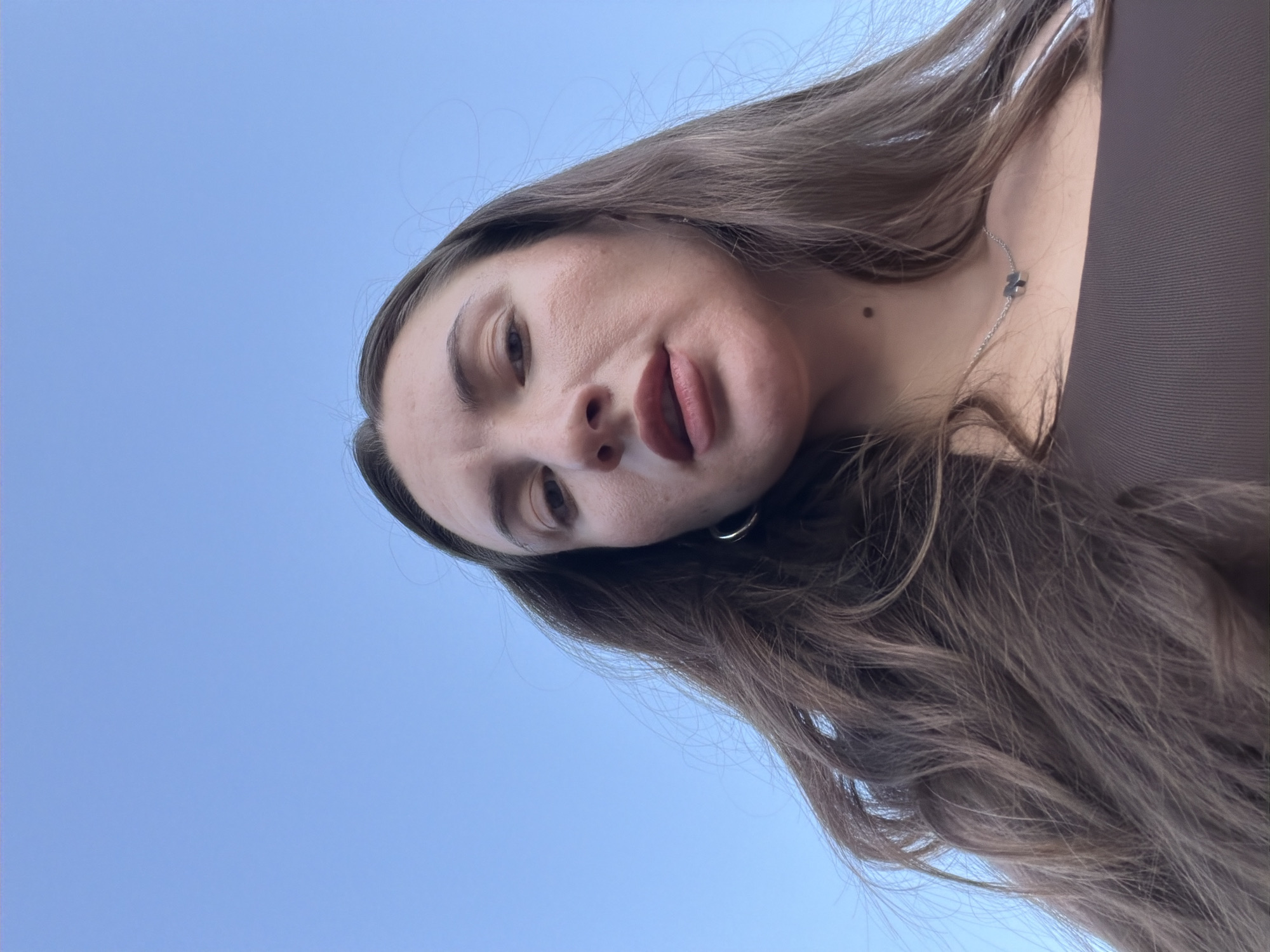 A low-angle, full-frame shot captures a woman looking upwards against a clear blue sky. Her face is angled towards the camera, with her mouth slightly open. She has long, dark brown hair that cascades around her shoulders and chest. She is wearing a brown tank top and a delicate silver necklace with a small pendant. A subtle mole is visible on her neck, just below her jawline. The background consists entirely of the bright blue sky, suggesting an outdoor setting. The image is devoid of any identifiable landmarks of Sharm El Sheikh, Egypt, or any other specific geographical indicators.