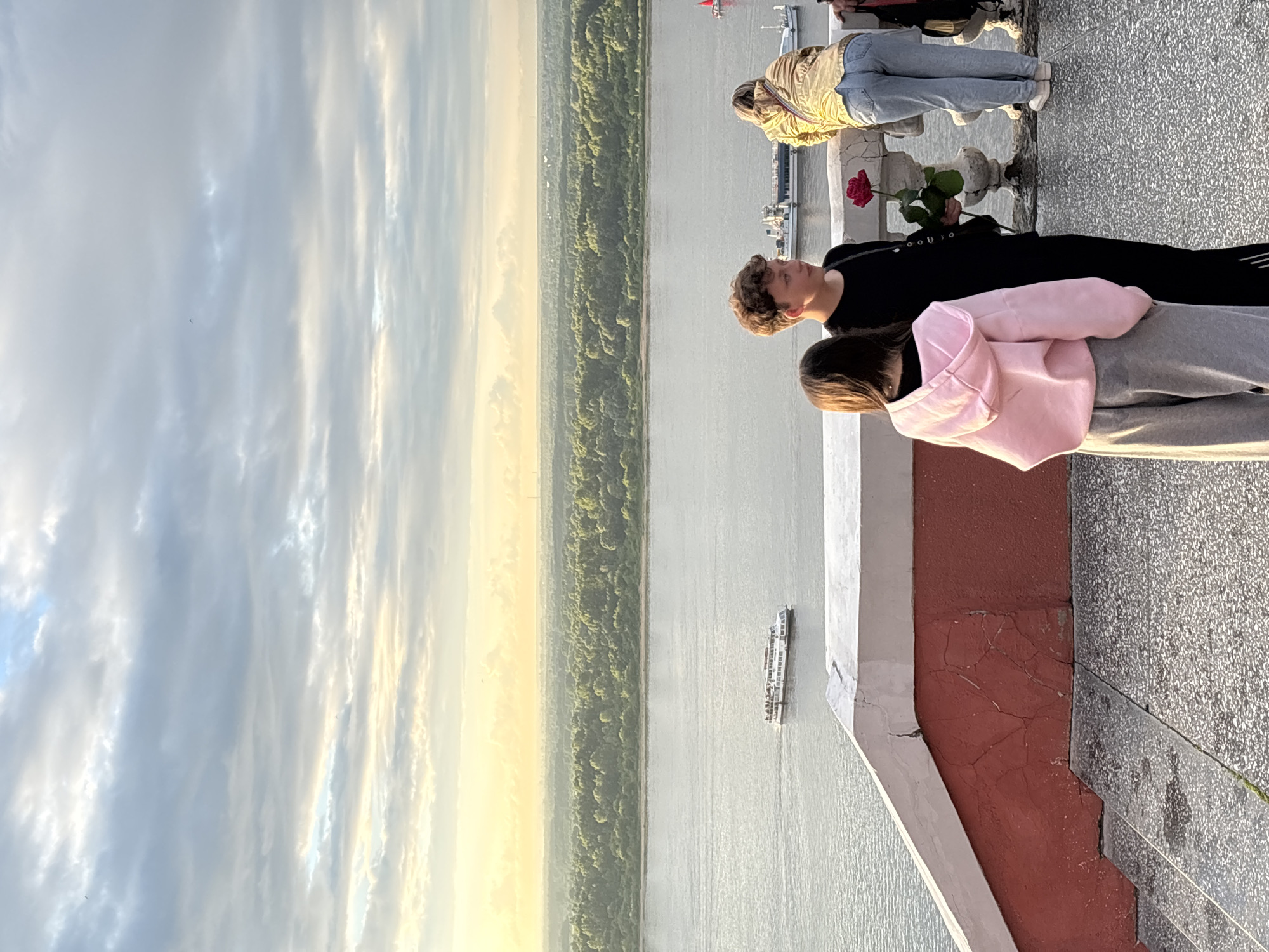 The image depicts a scenic overlook in Nizhny Novgorod, Russia, with a wide river in the foreground and a lush green forest on the opposite bank. In the distance, the sky is filled with dramatic clouds, illuminated by a soft, golden light.
In the foreground, three individuals are visible. A young woman wearing a pink hoodie and grey sweatpants stands with her back to the camera, looking out at the view. To her right, a young man in a black shirt and pants holds a single red rose. He appears to be looking towards the woman, and they are positioned near a white balustrade overlooking the river. Further to the right, another individual, possibly a woman, leans on the balustrade, facing away from the camera and wearing a gold jacket and blue jeans.
A white ferry boat with multiple decks is seen navigating the river, heading towards the left side of the frame. The water is calm and reflects the light from the sky. The overlook itself is characterized by a textured stone ground and a red and white painted railing, with visible cracks on the red portion. Stairs are also visible leading down to the left. The overall atmosphere suggests a peaceful moment, perhaps at sunset, enjoyed by the visitors to this vantage point.