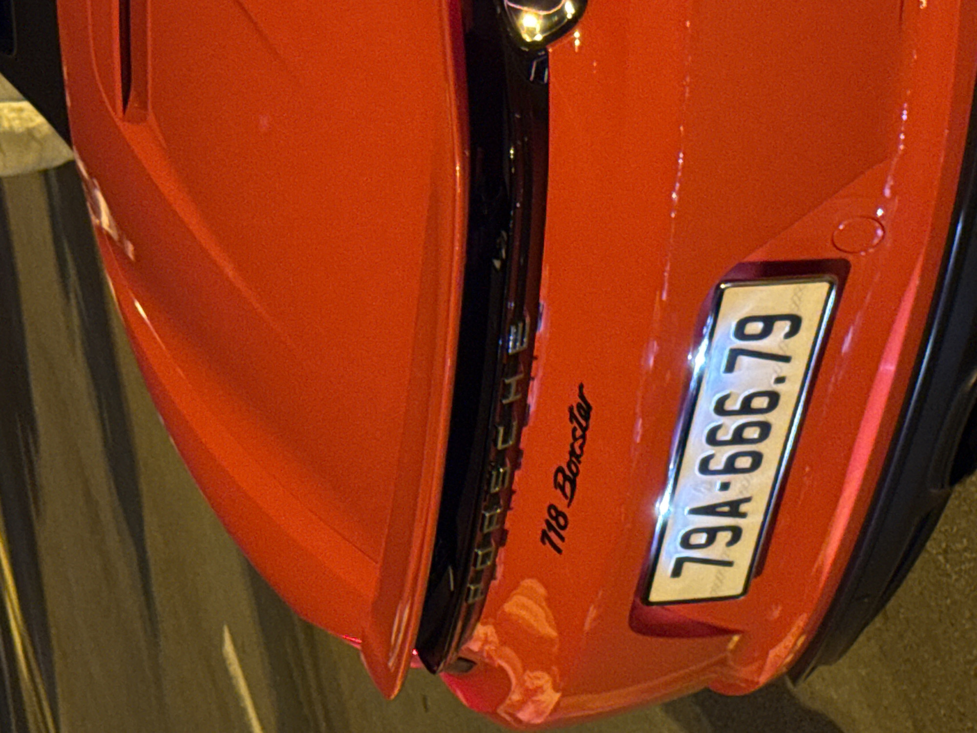 The image shows the rear of a red Porsche 718 Boxster parked on a street at night. The car's license plate reads "79A-666.79". The street is paved with asphalt and has yellow lane markings. A blurred figure can be seen in the upper right corner of the image. The scene is likely in Nha Trang, Vietnam, as indicated by the license plate format.