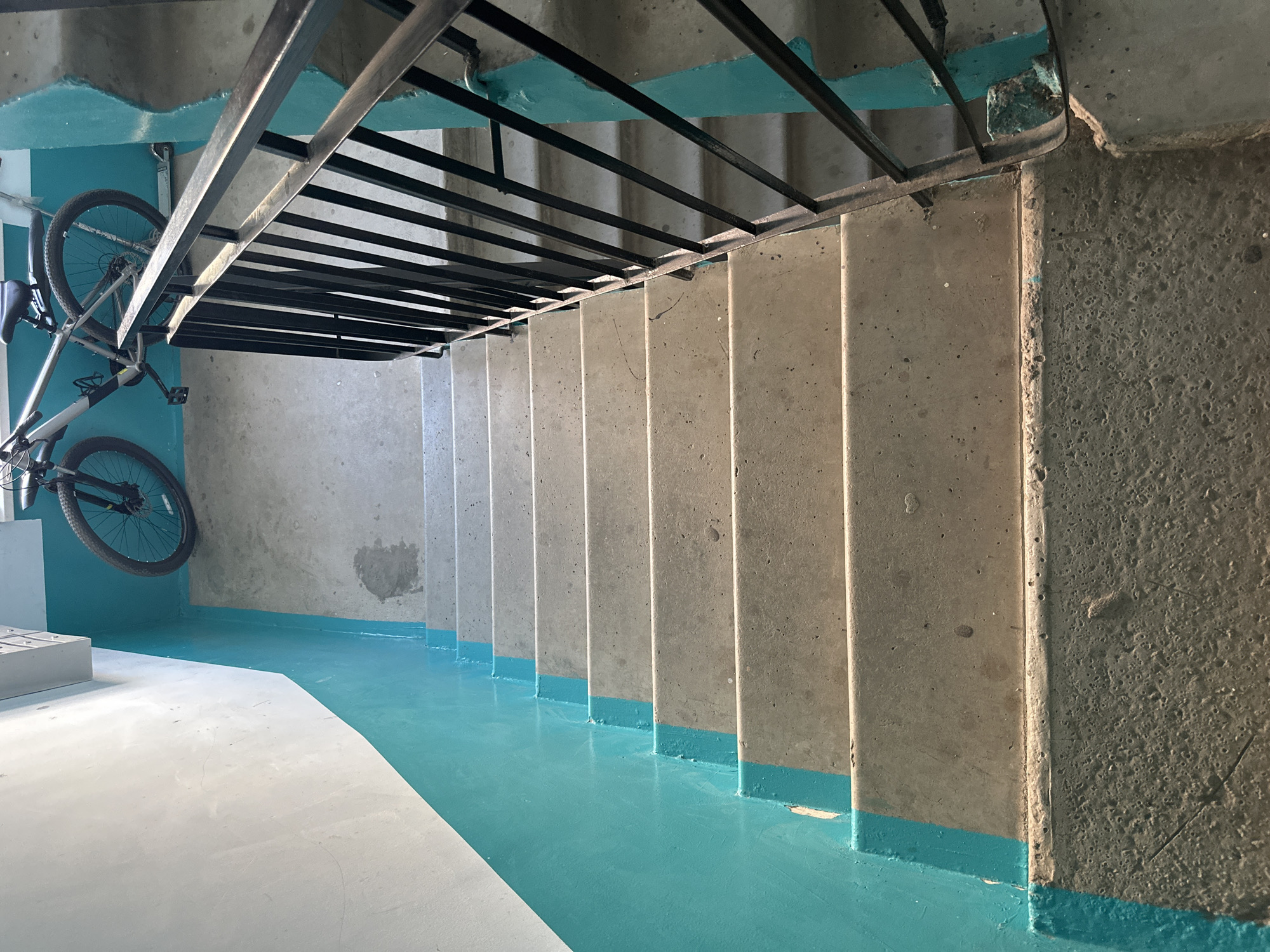 Here is a description of the image:
The image shows a set of concrete stairs leading downwards. The stairs are painted with a teal or turquoise color at the base of each step and on the adjacent floor. A black metal structure, likely a railing or some type of framework, runs along the ceiling above the stairs. A bicycle is mounted on the wall to the left of the stairs. The setting appears to be an interior stairwell, possibly in a building like an apartment complex or industrial space. The lighting is uneven, casting shadows across the stairs. The concrete appears worn and textured. The general location is not obvious from the context of the image.