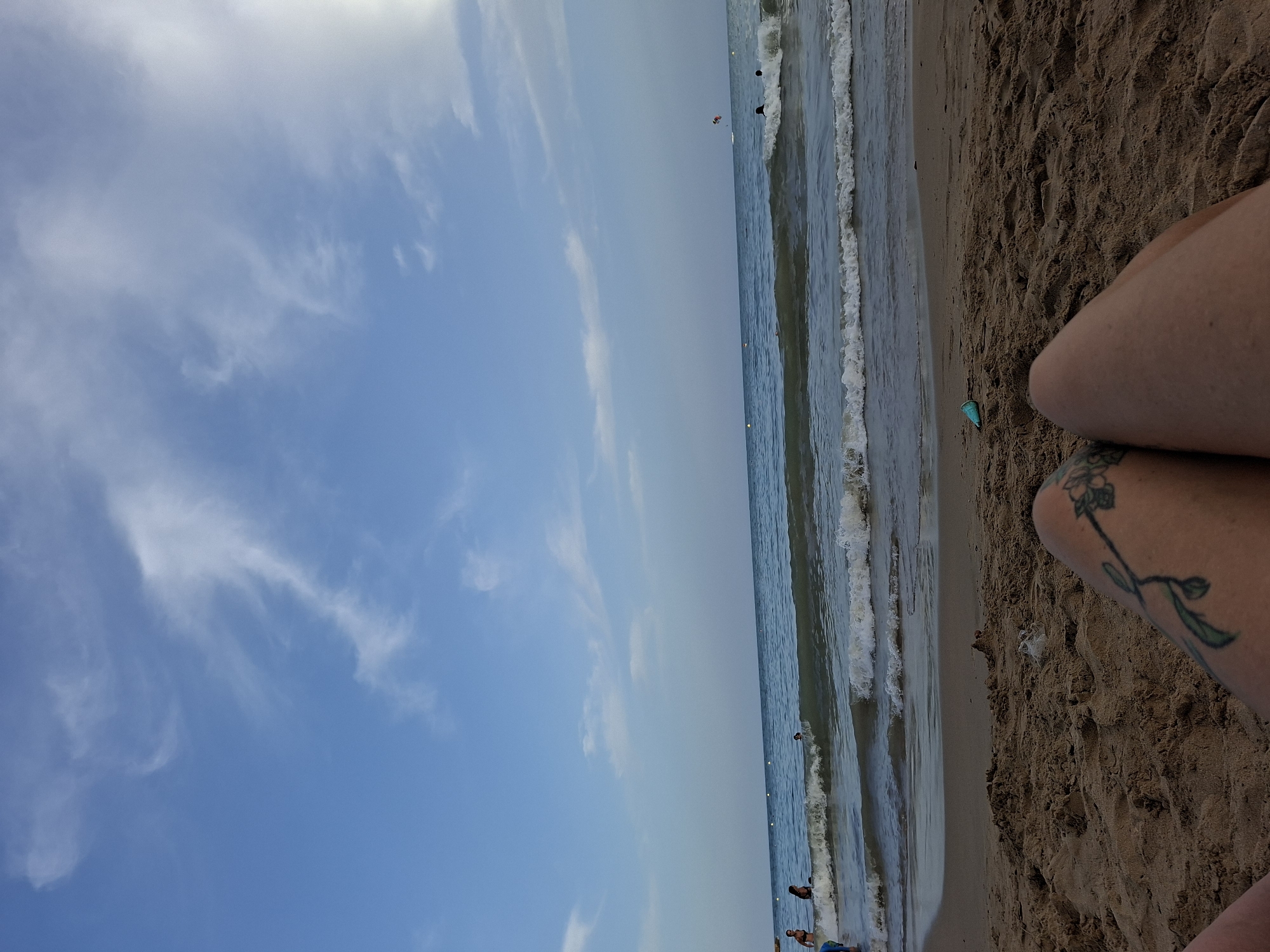The image depicts a beach scene on a clear day, with a view of the Mediterranean Sea. The foreground is dominated by wet sand, with the legs of a person visible, showing a tattoo on the left thigh. In the midground, gentle waves are breaking on the shore. Further out in the sea, several people are swimming or wading. In the distance, the calm sea stretches towards the horizon under a partly cloudy sky with wispy white clouds. A paraglider is visible in the far distance, adding a touch of activity to the expansive ocean view. The scene is likely set on one of Torrevieja's beaches, given the characteristic blue waters of the Mediterranean and the sandy coastline.