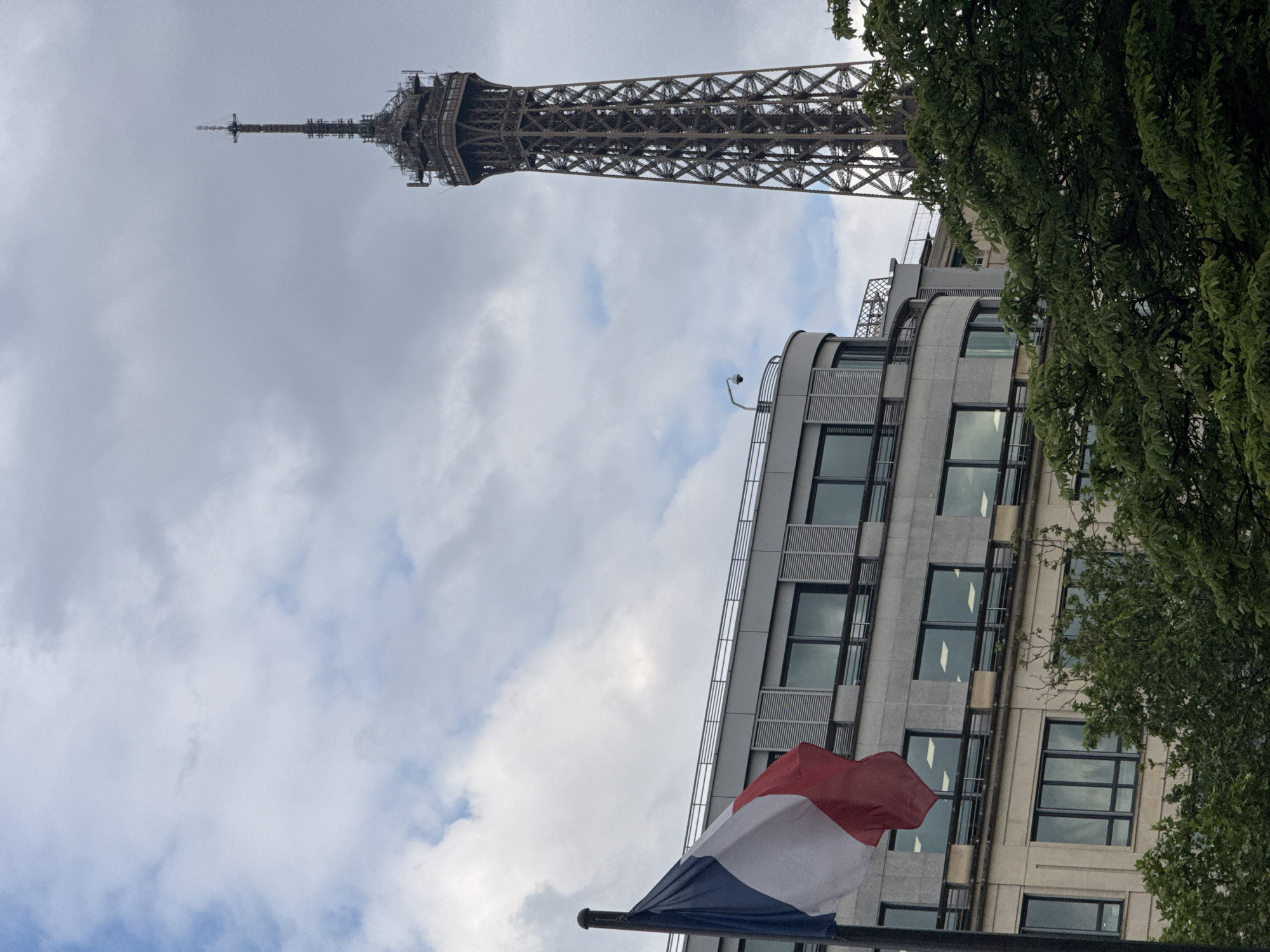 The image depicts the Eiffel Tower in Paris, France, angled upwards towards a cloudy sky. A modern building with multiple stories and windows is positioned adjacent to the tower, partially obscured by foliage on the right. The French flag is visible in the foreground, indicating a governmental or official location. No people are visible in the scene.