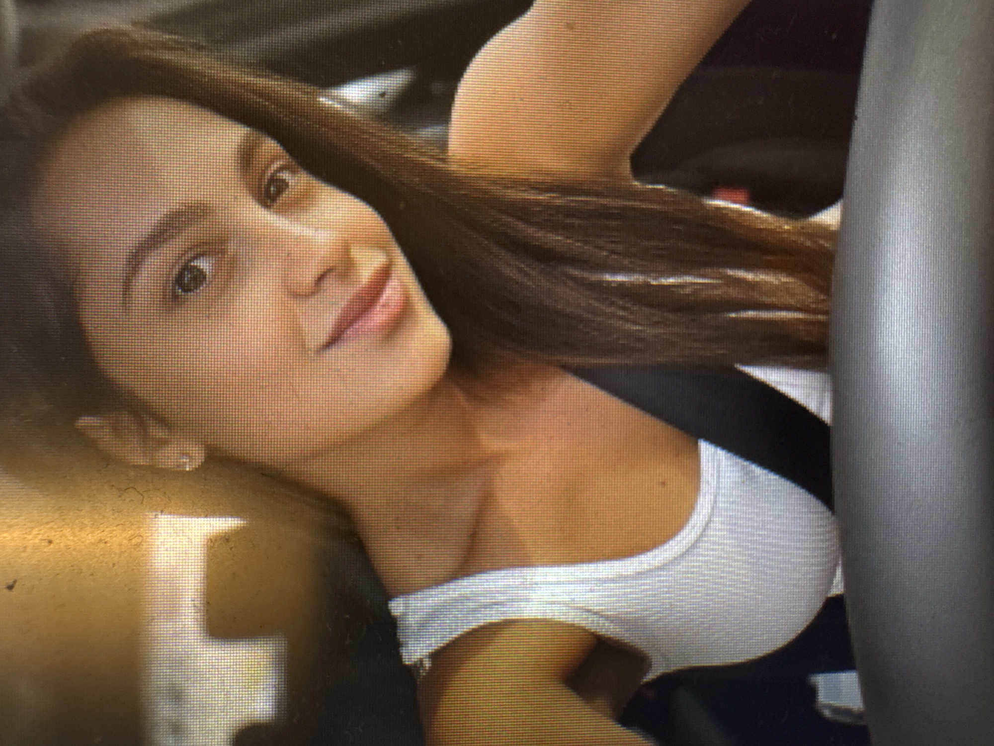 A close-up, head-and-shoulders portrait shows a young woman with long, dark brown hair and light brown eyes. She is wearing a white tank top and a black seatbelt. She is looking towards the camera with a slight smile. The background is blurry and appears to be the interior of a car, with some golden light suggesting daylight. There is no discernible detail within the image that would place this scene in Murcia, Spain.