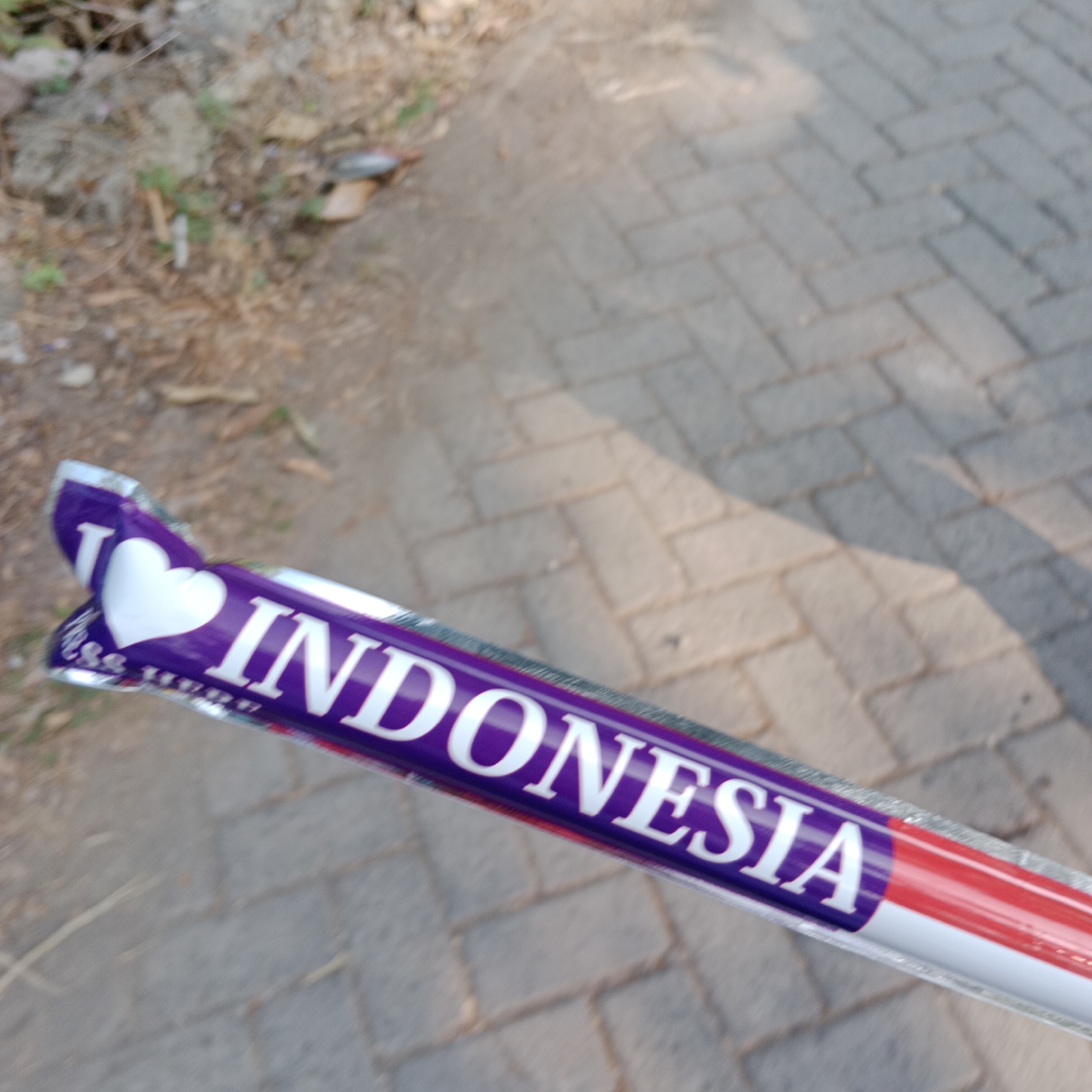 A hand is holding a purple celebratory noisemaker with a white heart and "INDONESIA" printed in white letters. The noisemaker is partially wrapped in silver foil. The background shows a brick pathway with sunlight casting shadows. Dry leaves and dirt are visible to the left of the pathway. The image is unfocused and was taken outdoors.