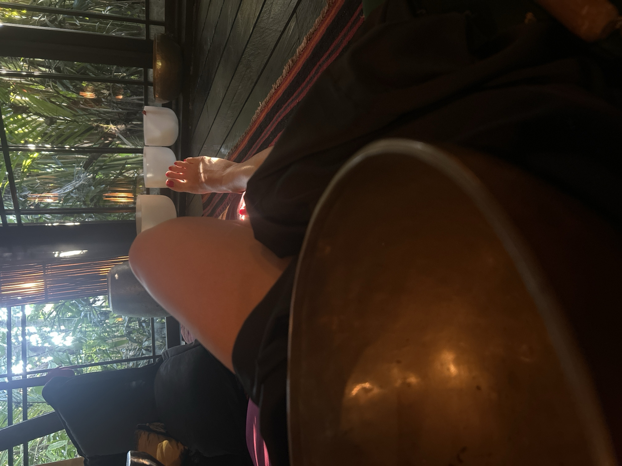 The image depicts a person's legs and feet resting on a wooden floor covered with a striped rug. A large, golden singing bowl is prominently featured in the foreground. In the background, through a large window or opening, lush green foliage and sunlight are visible. The setting appears to be an indoor space with natural light, possibly a relaxation or meditation room. There is no clear indication of the city of Kediri, Indonesia, within the image itself.
