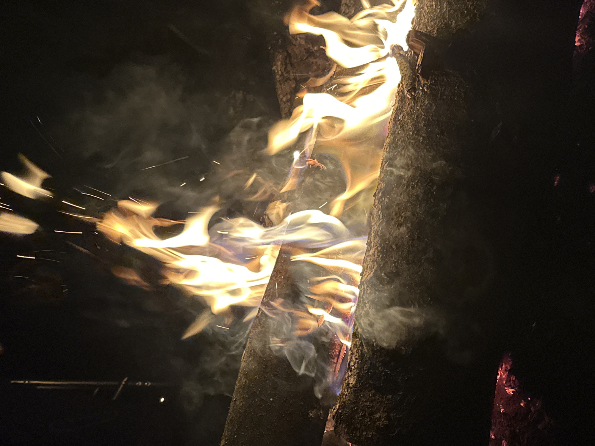 The image depicts a fire burning between two large logs in a dark environment. Sparks and smoke are visible around the flames. The logs appear to be charred. The location is not discernible from the image.
