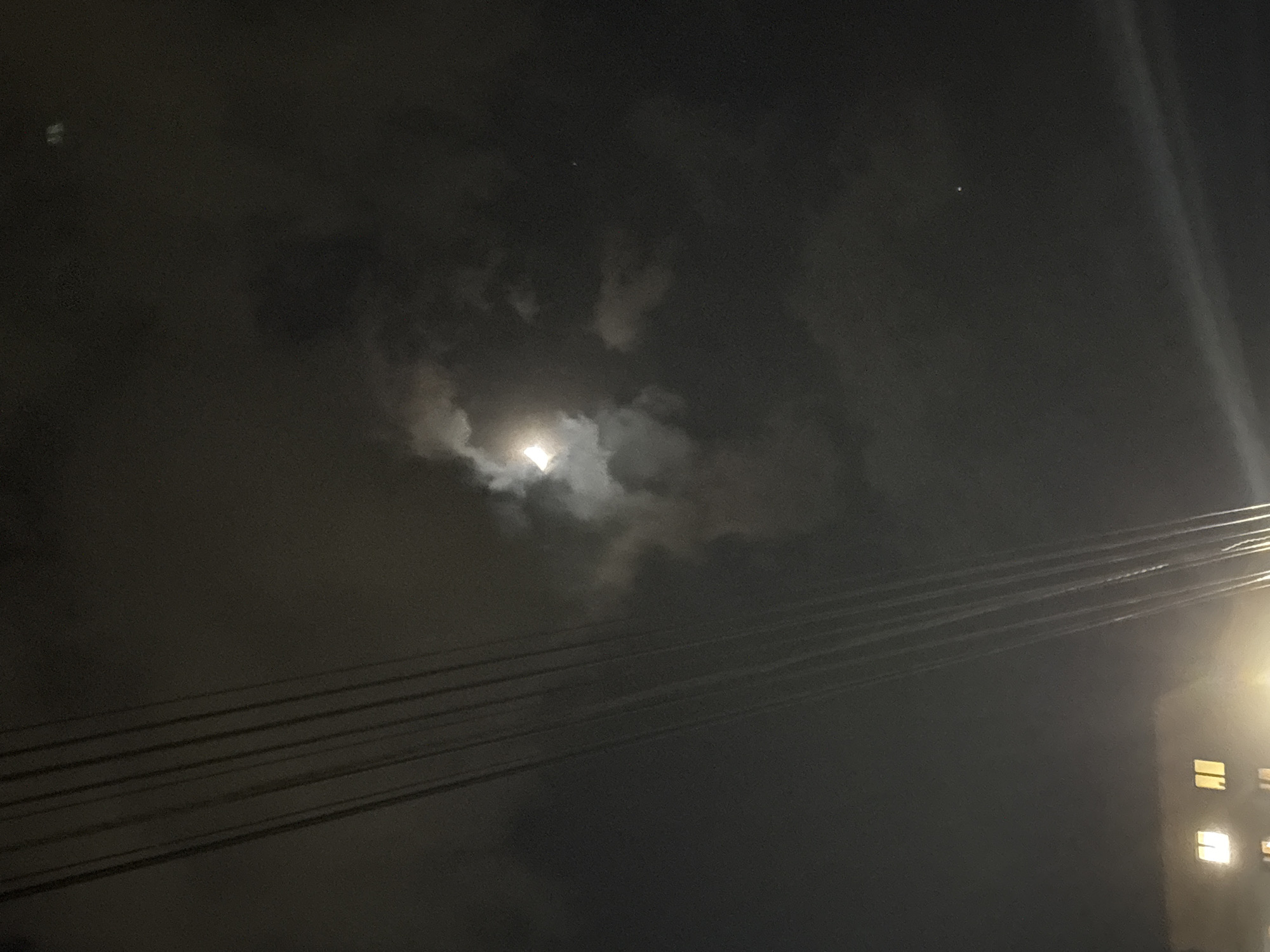 This media file presents a nighttime urban landscape, likely in Yerevan, Armenia. The dominant feature is a dark, cloudy sky, with a bright, partially visible moon glowing centrally through a break in the dense, grey clouds. The moon acts as a primary light source, illuminating the edges of the surrounding clouds.
Stretching diagonally across the lower right portion of the frame are several dark, parallel power lines or cables. These lines appear to connect towards a visible segment of a building on the far right. This building features a few brightly lit windows, which stand out against its dark facade and contribute to the urban setting.
In the upper left quadrant of the sky, two very faint, small white dots are discernible, consistent with distant stars or aerial lights. The image captures a serene and atmospheric night scene, characterized by the interplay of natural moonlight filtering through clouds and artificial light emanating from the urban environment. There is no visible text in the image.