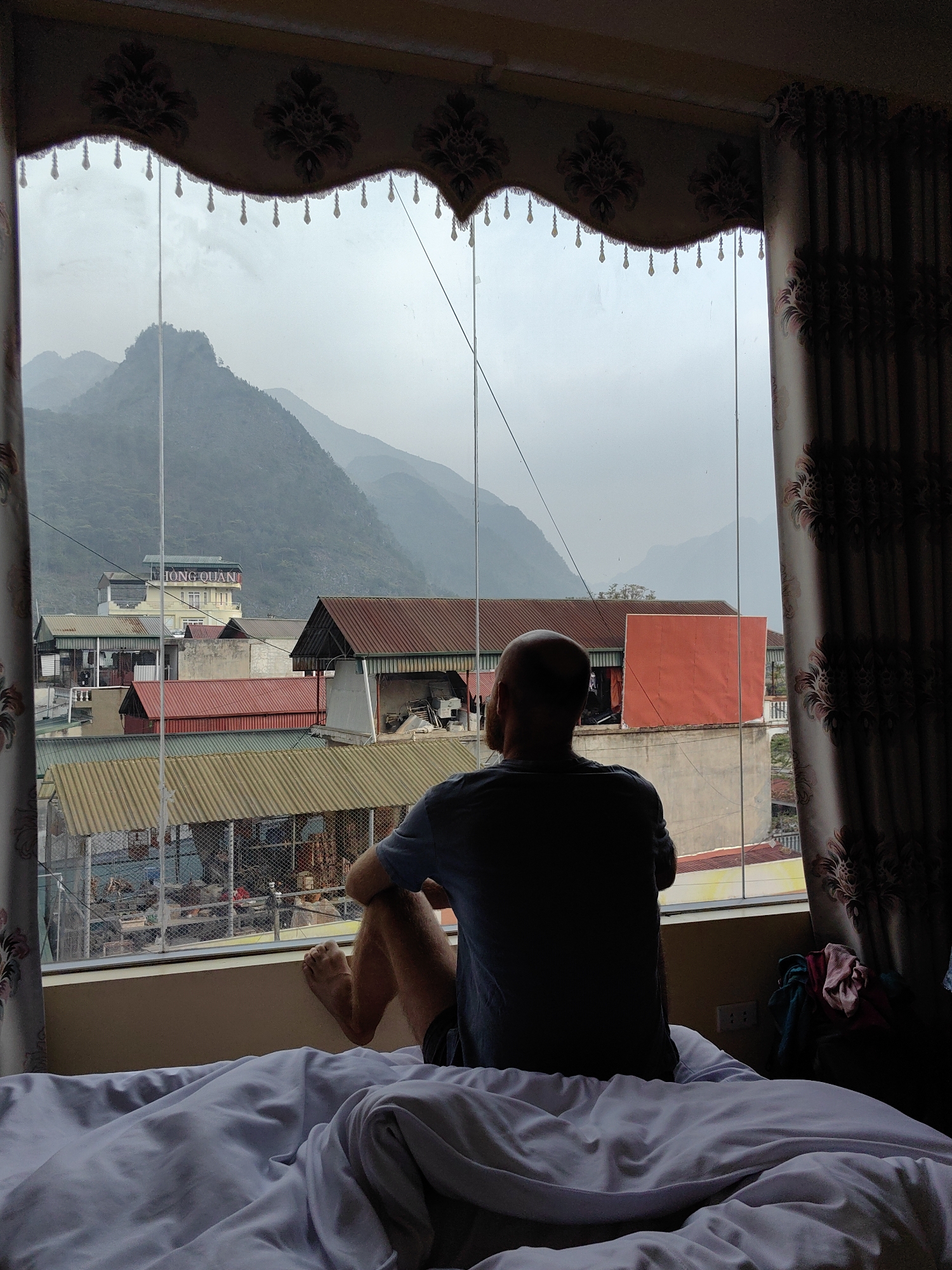 A person sits on a bed, looking out a window in a building in the Mèo Vạc Commune, Vietnam. The person is facing away from the camera, with their back visible. A blue t-shirt is worn, and legs are bent at the knees. The bed has white sheets and is rumpled. The window looks out onto a scene of buildings, rooftops, and mountains in the background. There are curtains with a patterned design framing the window. Outside, the sky is overcast. The text "HONG QUAN" is visible on a building in the distance.