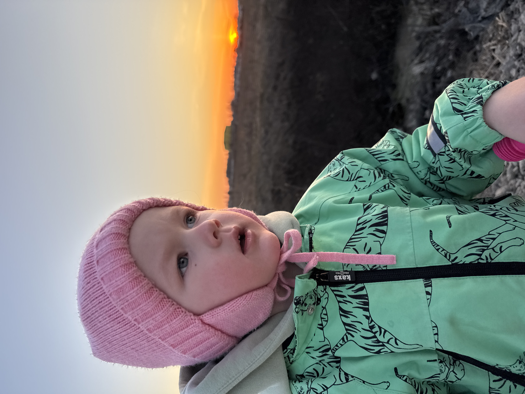 A toddler wearing a pink knitted hat and a green jacket with a tiger print is the main subject of the image. The child is looking upwards with an open mouth. The background features a sky gradient transitioning from light blue to orange, indicating either sunrise or sunset. Below the sky, there is a dark area, likely representing land or vegetation. The picture is taken outdoors, possibly in a park or a field. It's difficult to determine the location specifically as Горный, Russia from this picture alone.