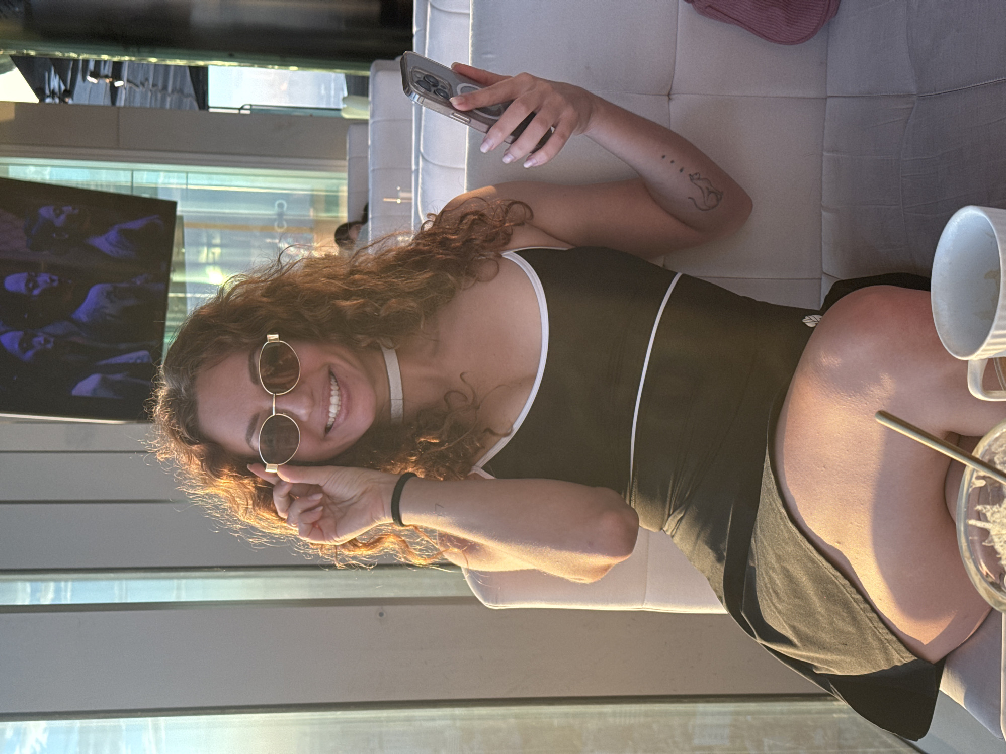A young woman with curly brown hair and sunglasses is sitting on a white couch, holding her phone and adjusting her sunglasses. She is wearing a black dress with white trim. She has a tattoo of a cat on her arm. In the background, there are large windows and a television screen showing images of people. There are also other people sitting on couches in the background. The lighting suggests it is late afternoon or early evening. The environment appears to be a modern lounge or restaurant. There is no indication that this scene is located in Hà Nội, Vietnam.